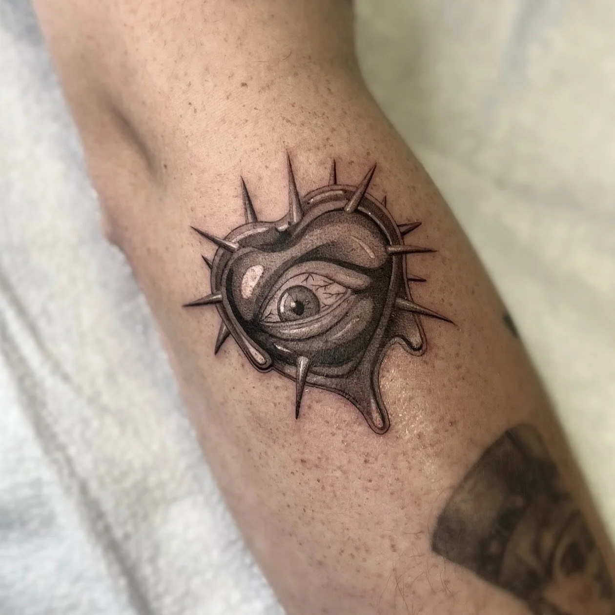 Tatouage d'un œil dans un cœur avec des épines autour, en nuances de gris, sur la peau d'une personne.