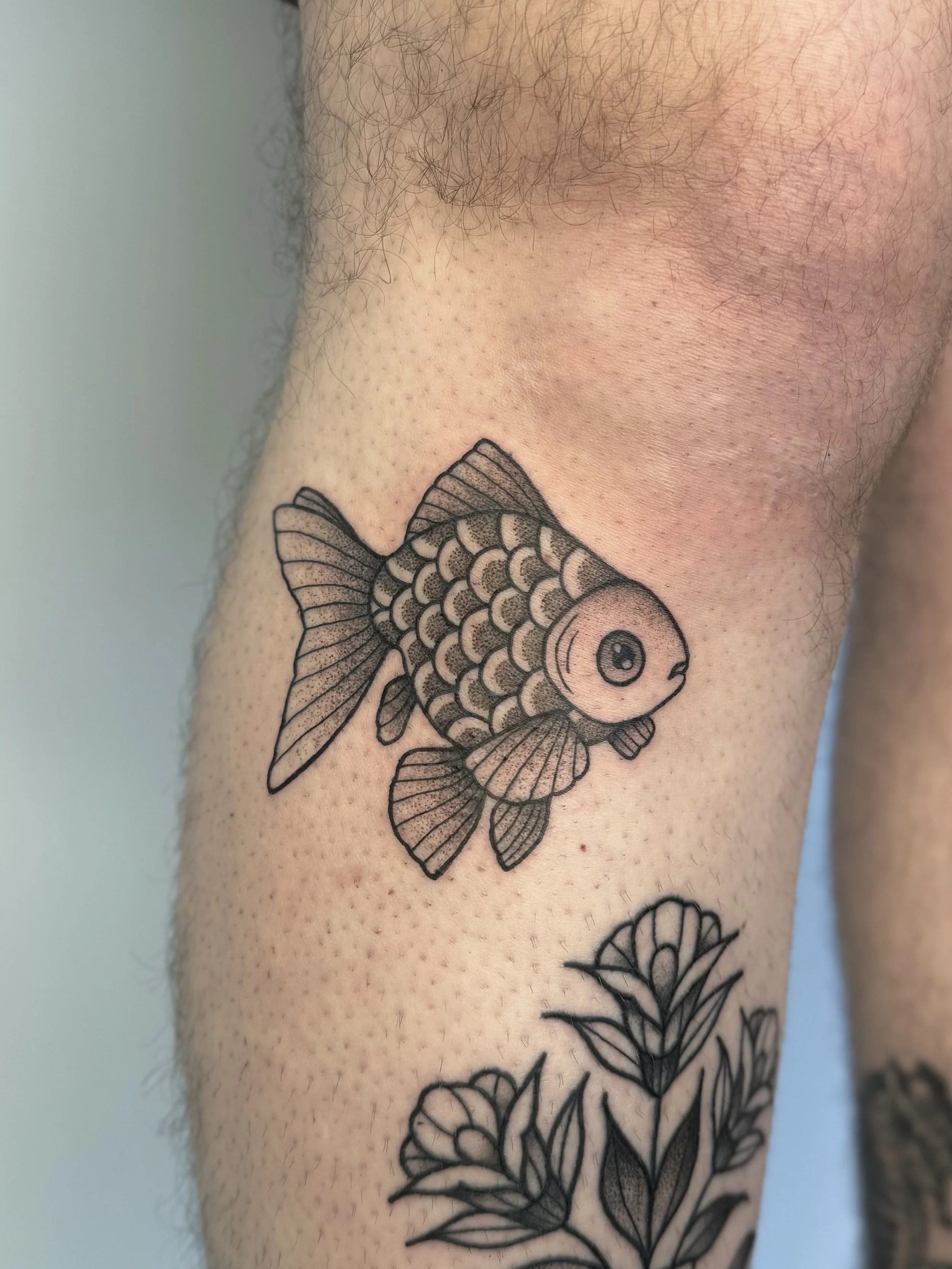 Tatouage en noir et blanc d’un poisson rouge sur la cuisse, avec des détails fins et des écailles, à côté de feuilles de plantes.