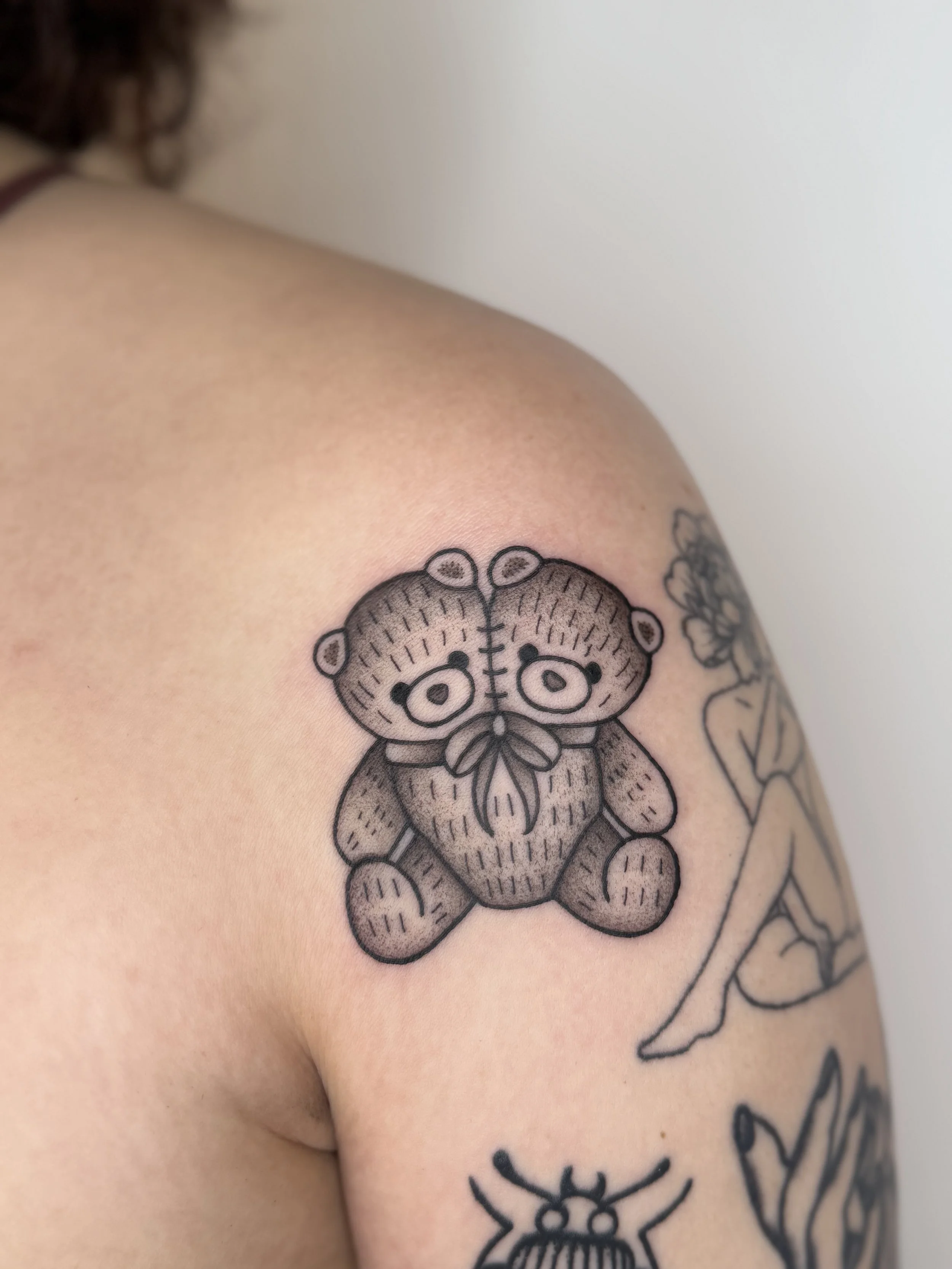 Tatouage d'un ours en peluche dessiné en noir sur l'épaule d'une personne, avec d'autres tatouages visibles à côté.