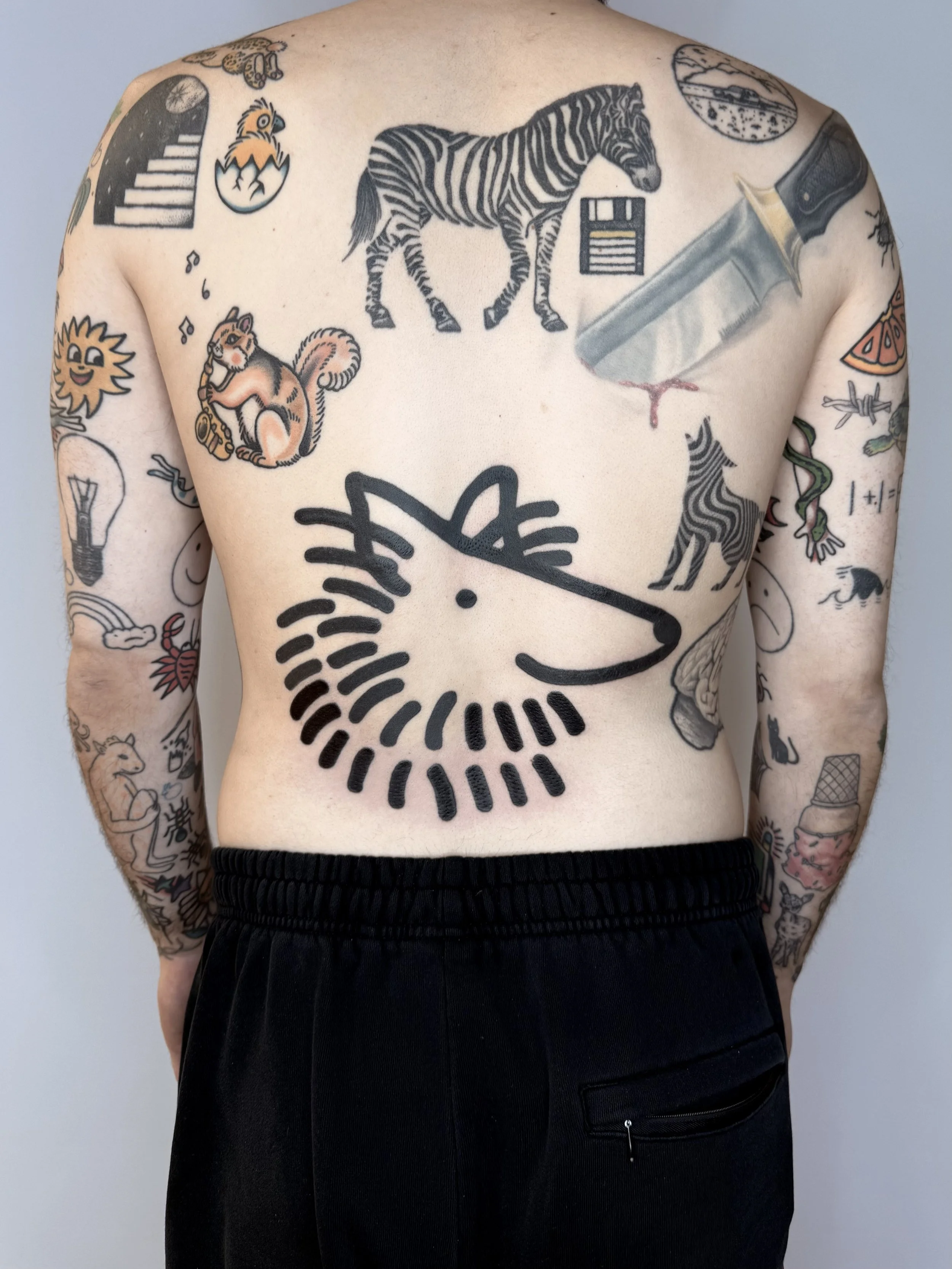 Tatouages colorés et noirs sur le torse et les bras d'une personne; dont un lion stylisé, un zèbre, un couteau avec du sang, un chat, une horloge, une guitare, un soleil souriant, un animal avec des cordes, des mélodies, un papillon, un dragon, une p