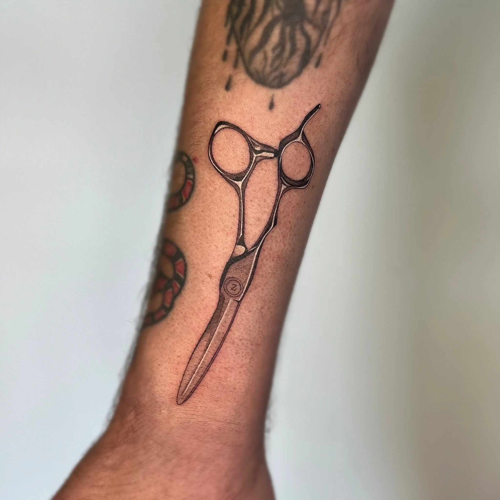 Tatouage d'une paire de ciseaux chirurgicales sur l'avant-bras avec d'autres tatouages partiellement visibles.