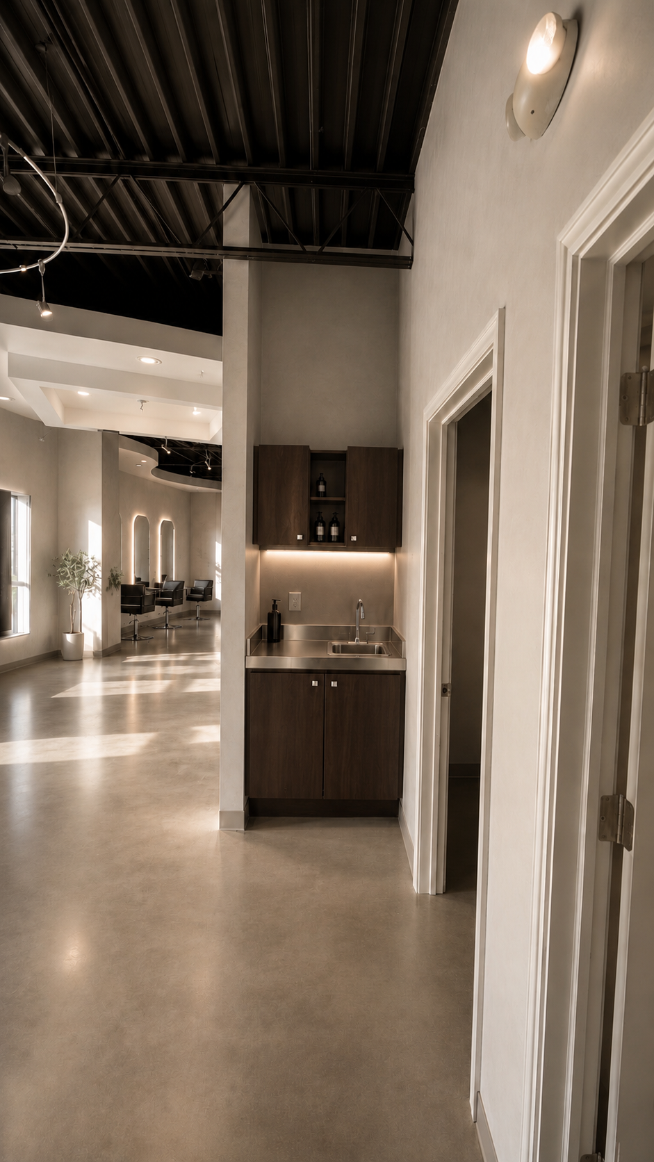 Interior of a modern room with a small stainless steel sink and dark wooden cabinetry adjacent to a doorway, with a large open area and seating in the background.