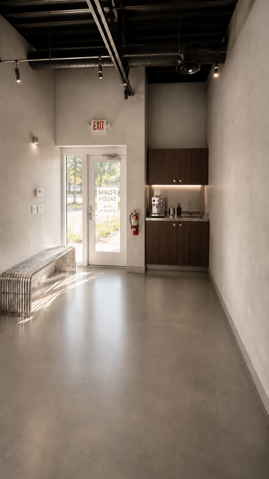 Minimalist interior with a light gray polished concrete floor, white walls, a metal bench, a glass door with a "Form Salon" sign, a red fire extinguisher, and a small kitchenette with dark wood cabinets, a coffee machine, and a sink, illuminated by c