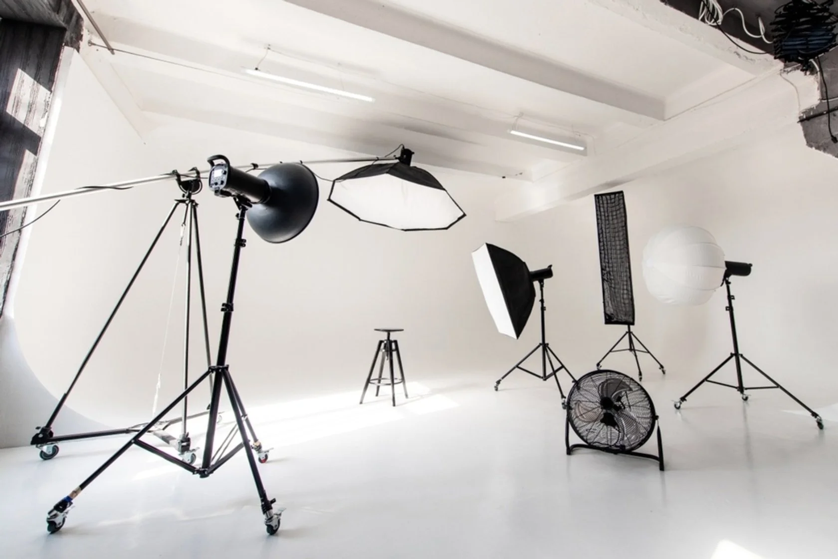 Photography studio with professional lighting equipment, including softbox lights, umbrella lights, a fan, and a white backdrop, all set up in a white room.
