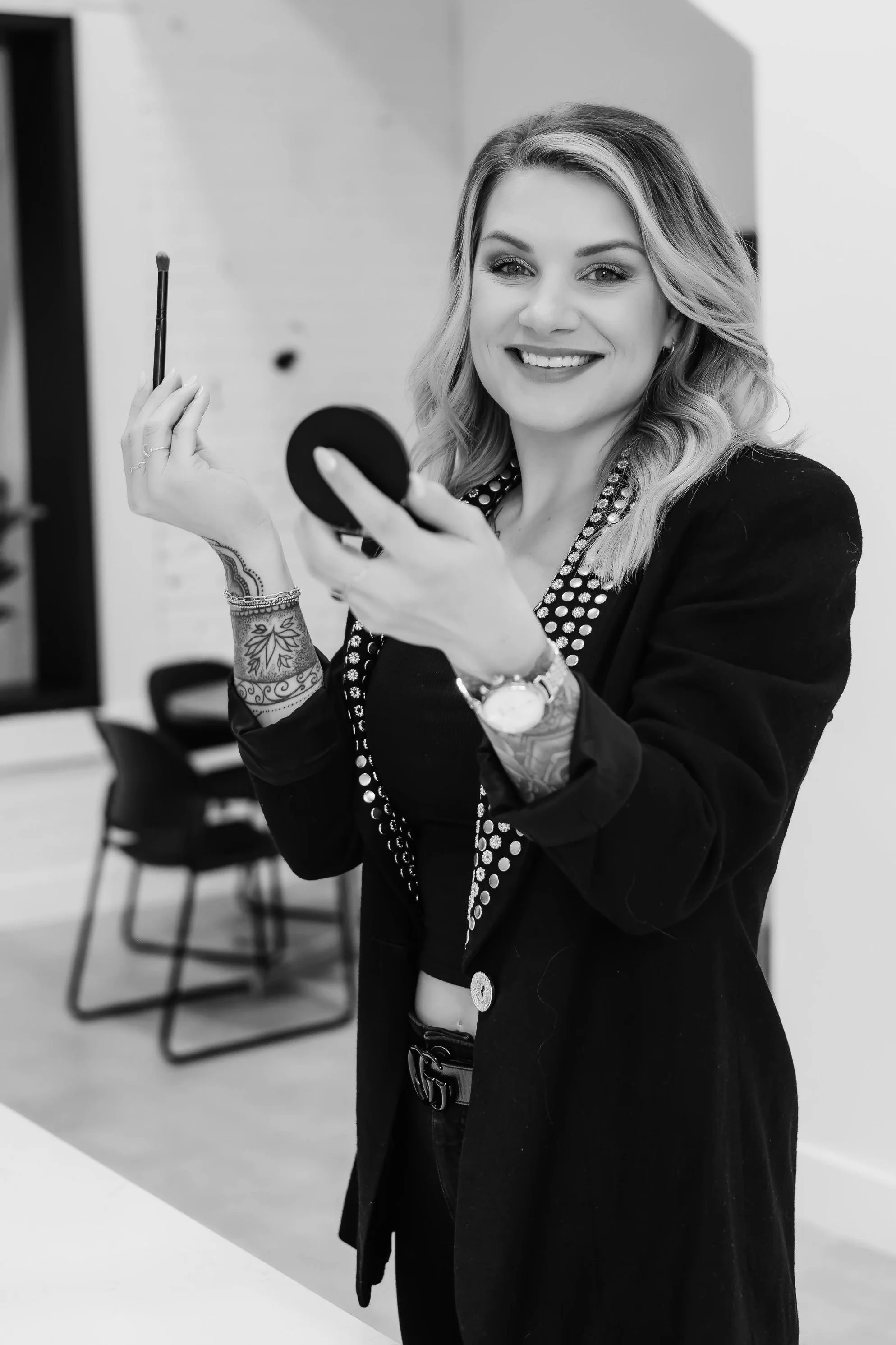 A woman with wavy hair and tattoos on her arm, holding a makeup brush and a compact mirror, smiling at the camera in a room with chairs and a table in the background.