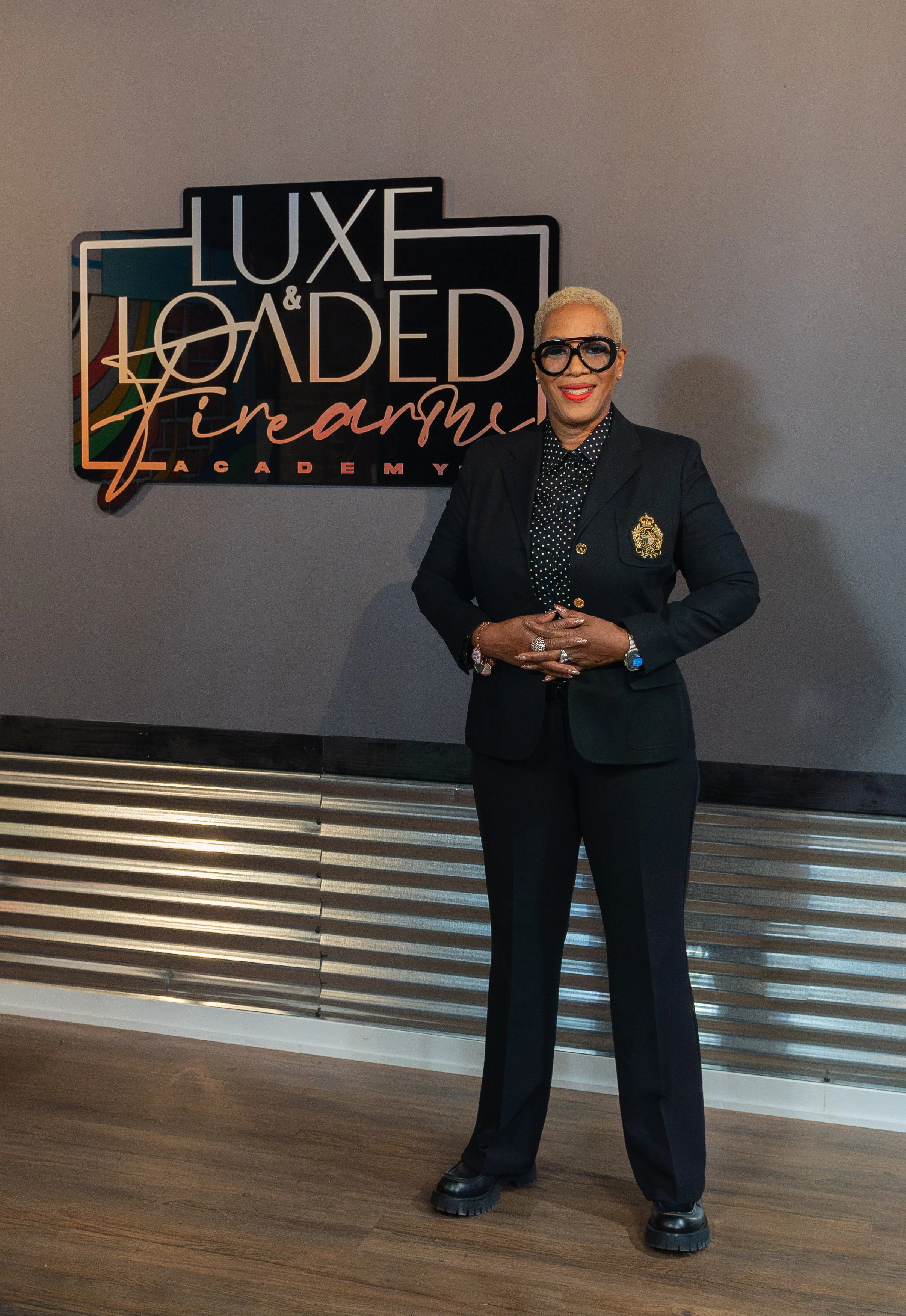 A woman in a black suit and large glasses standing in front of a sign that reads 'Luxe & Loaded Firearms Academy'