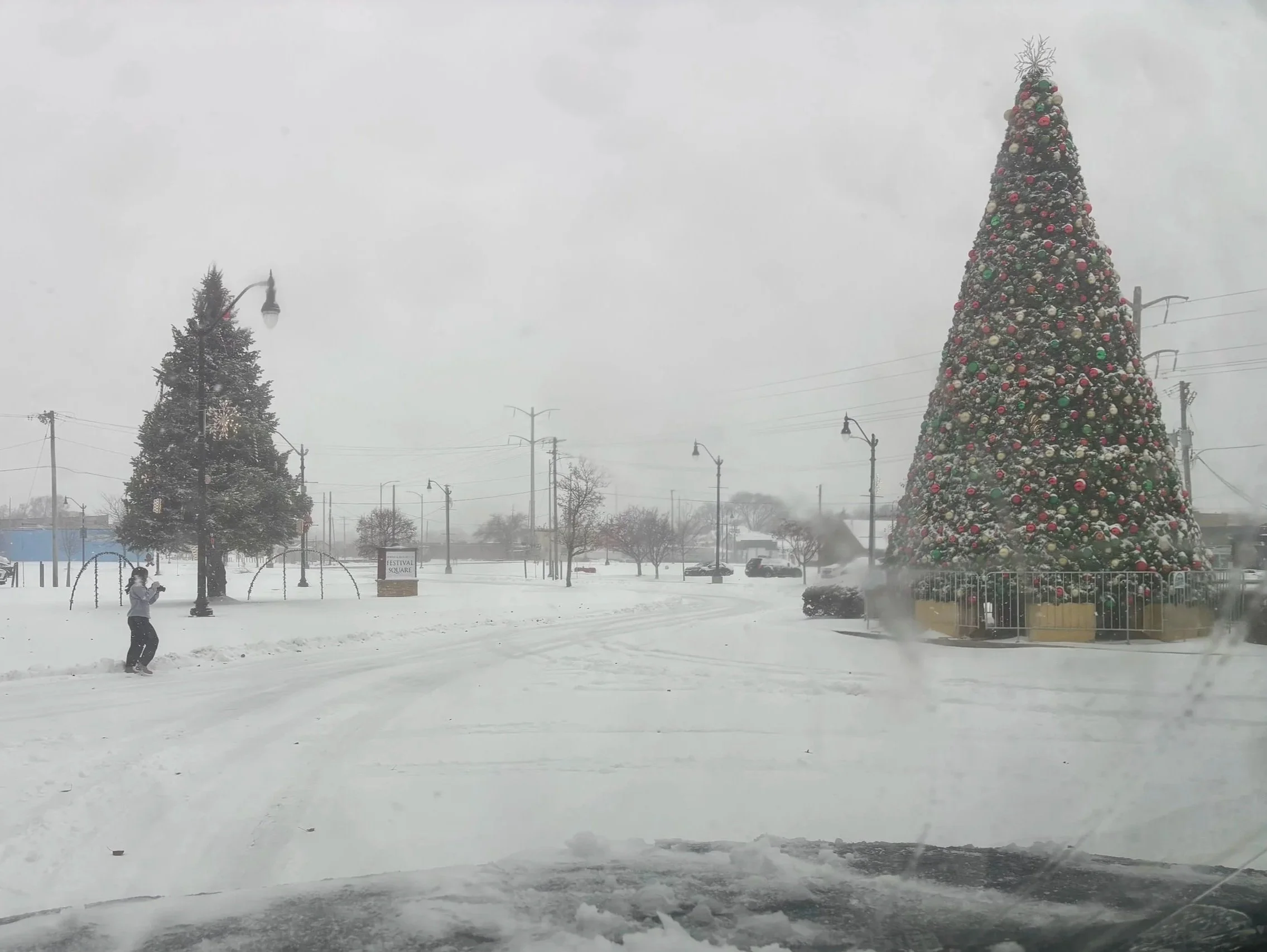 A large decorated Christmas tree stands in a snow-covered town square, surrounded by a white picket fence. Nearby, a person is playing in the snow. The scene is foggy with snow falling, and other trees and streetlights are visible in the background.