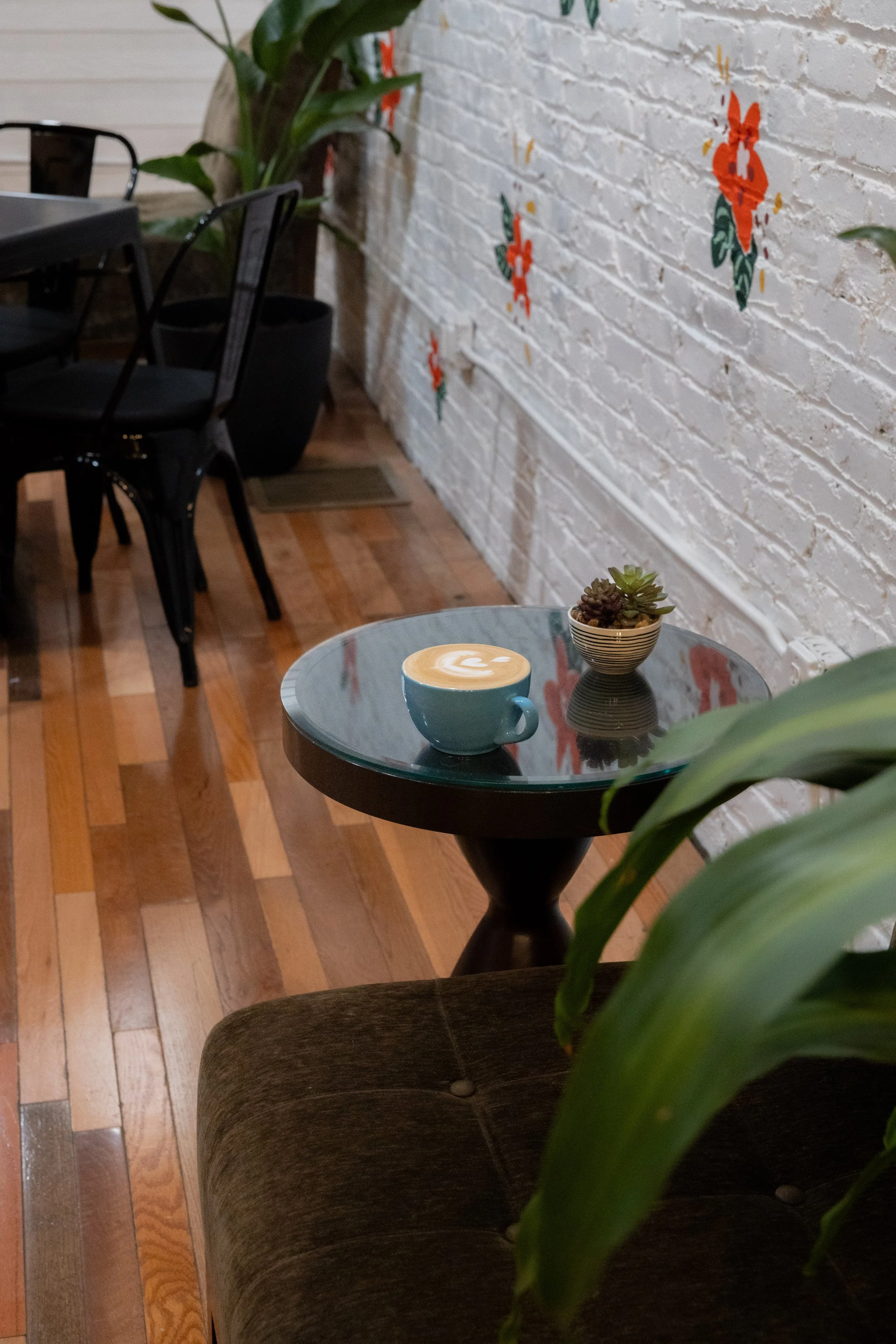 A cozy cafe interior with a small round glass-topped table holding a latte in a blue mug and a small pot of succulents. The background features a white brick wall decorated with stylized red and green floral artwork, and dark wooden flooring.