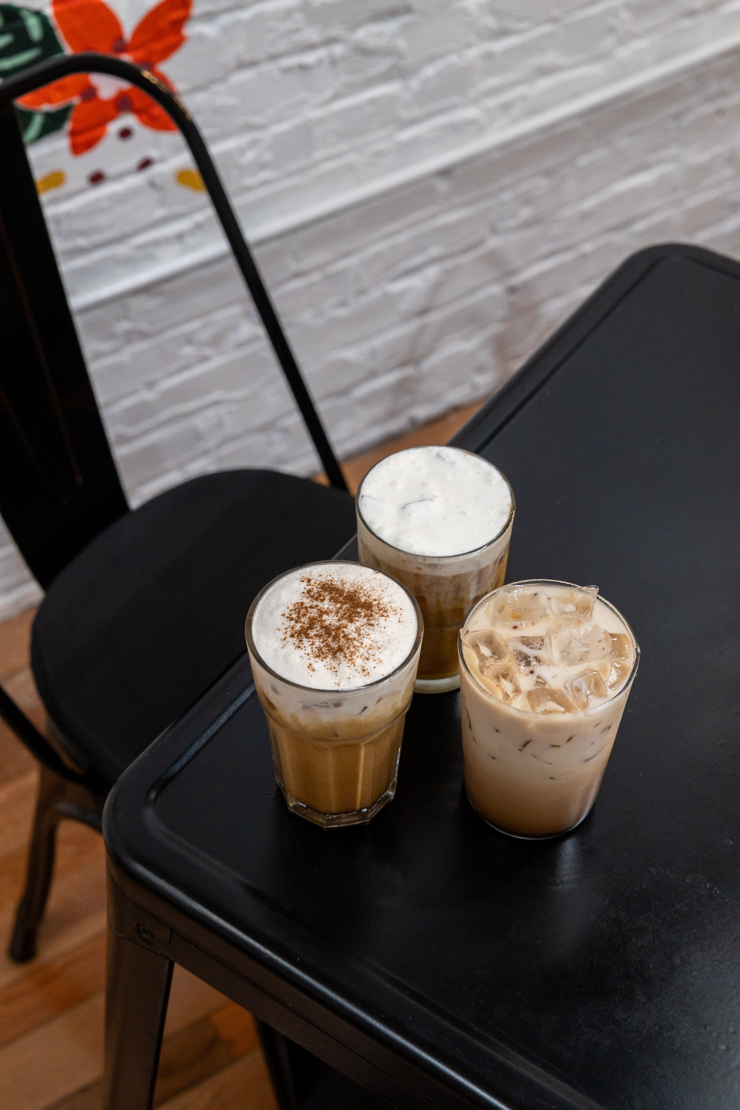 Three iced coffee drinks on a black table, with one topped with cinnamon, another with foam, and the third with ice cubes, in a cozy cafe setting with a white brick wall and black chair in the background.