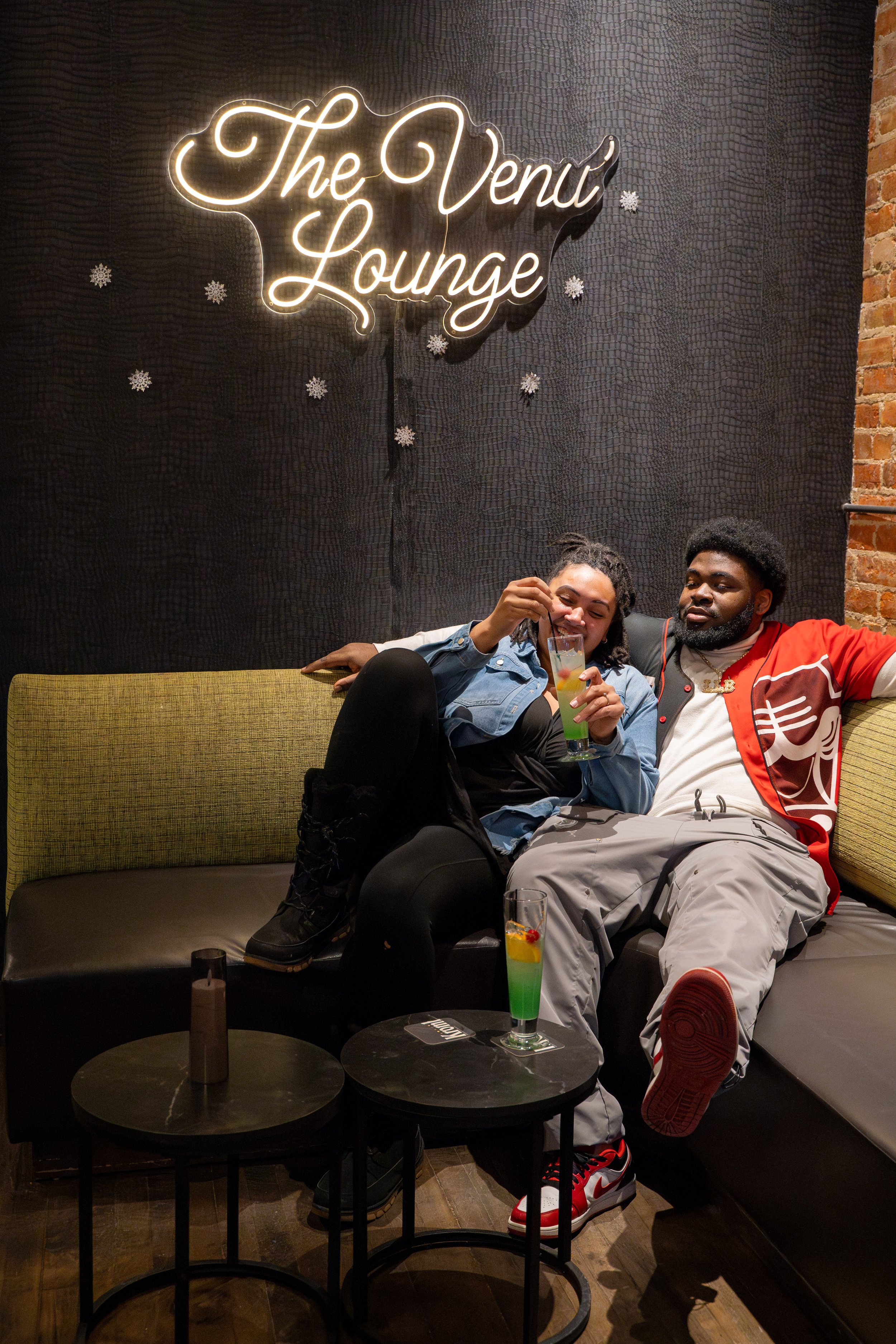 Two people sitting on a green-cushioned black sofa, smiling and enjoying drinks in a lounge with a sign that reads "The Venu Lounge" on the wall behind them. The woman is wearing a denim jacket and black clothing, while the man is wearing a red and w