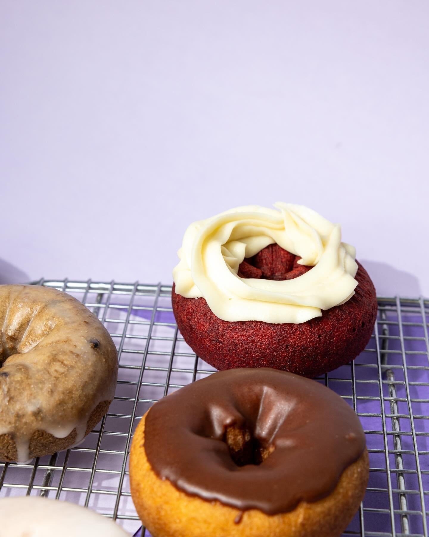 Three donuts on a cooling rack, with a purple background. One is a red velvet donut with white icing on top, another is a chocolate-glazed donut, and another appears to be a cinnamon or caramel-flavored donut.