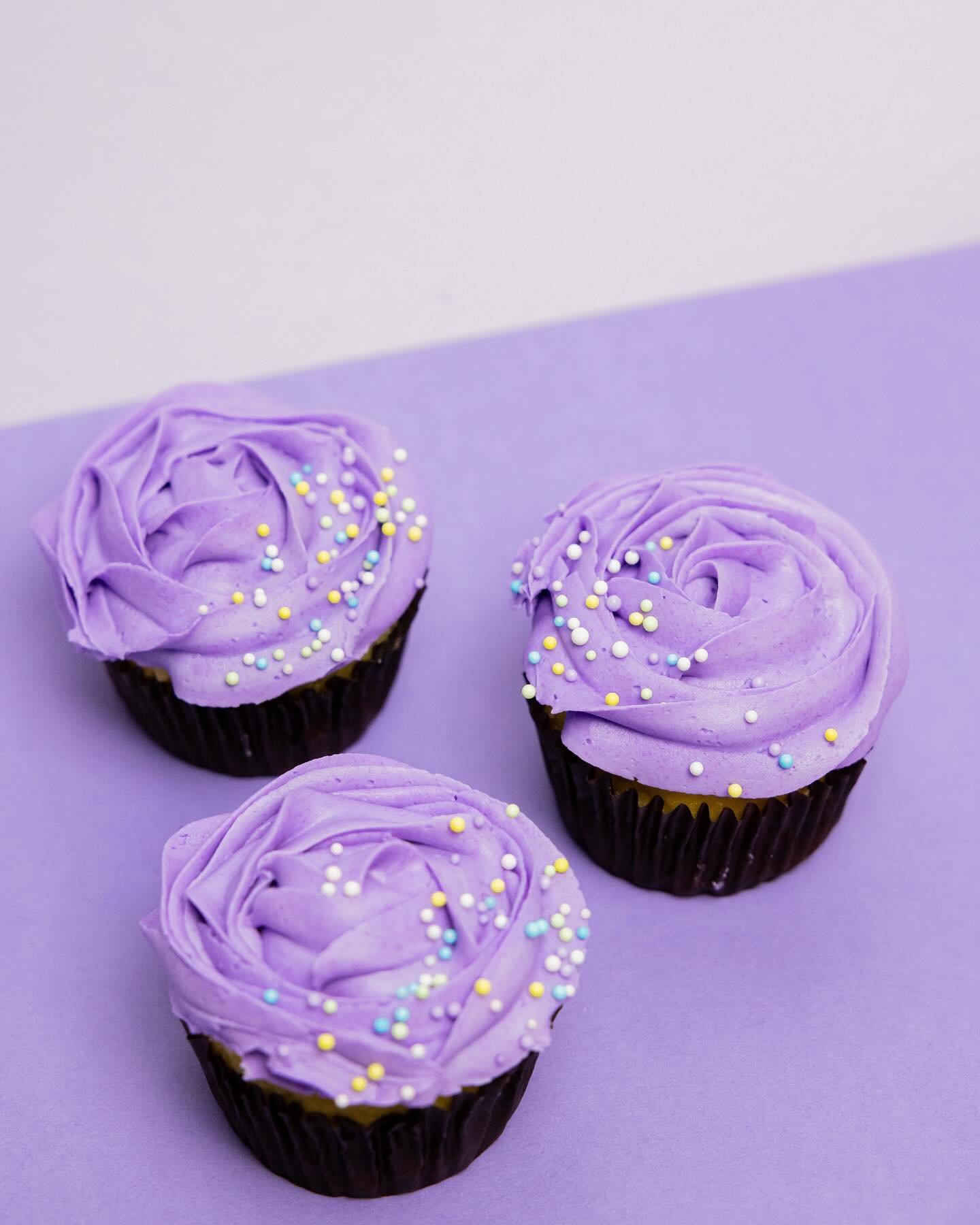 Three cupcakes with purple frosting and colorful sprinkles on a purple surface.