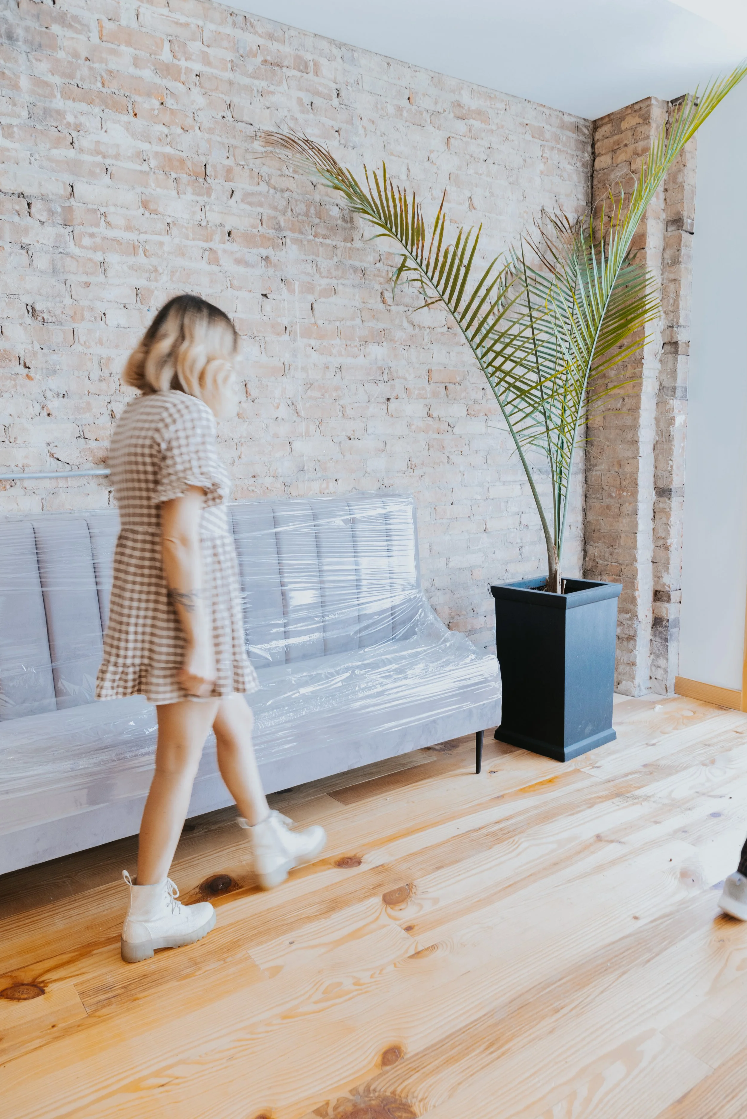 A woman with blonde hair wearing a brown and white checkered dress and white boots walking past a plastic-covered sofa in a room with brick wall and wooden floor and a tall potted plant.
