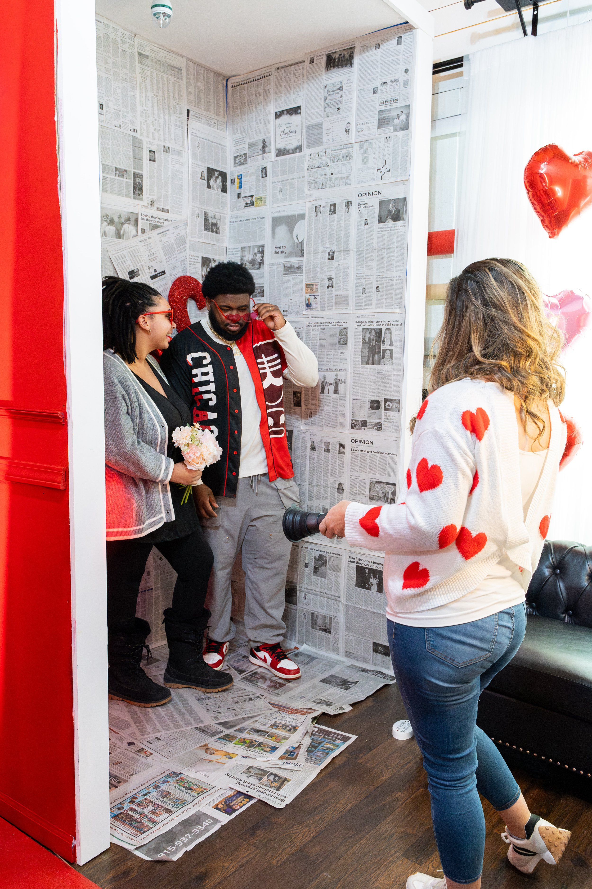 A woman with curly hair wearing a white sweater with red hearts takes a photo of a couple in a small photo booth covered with newspapers. The woman holding a camera faces the couple who are standing in the booth. The woman in the couple is holding a 