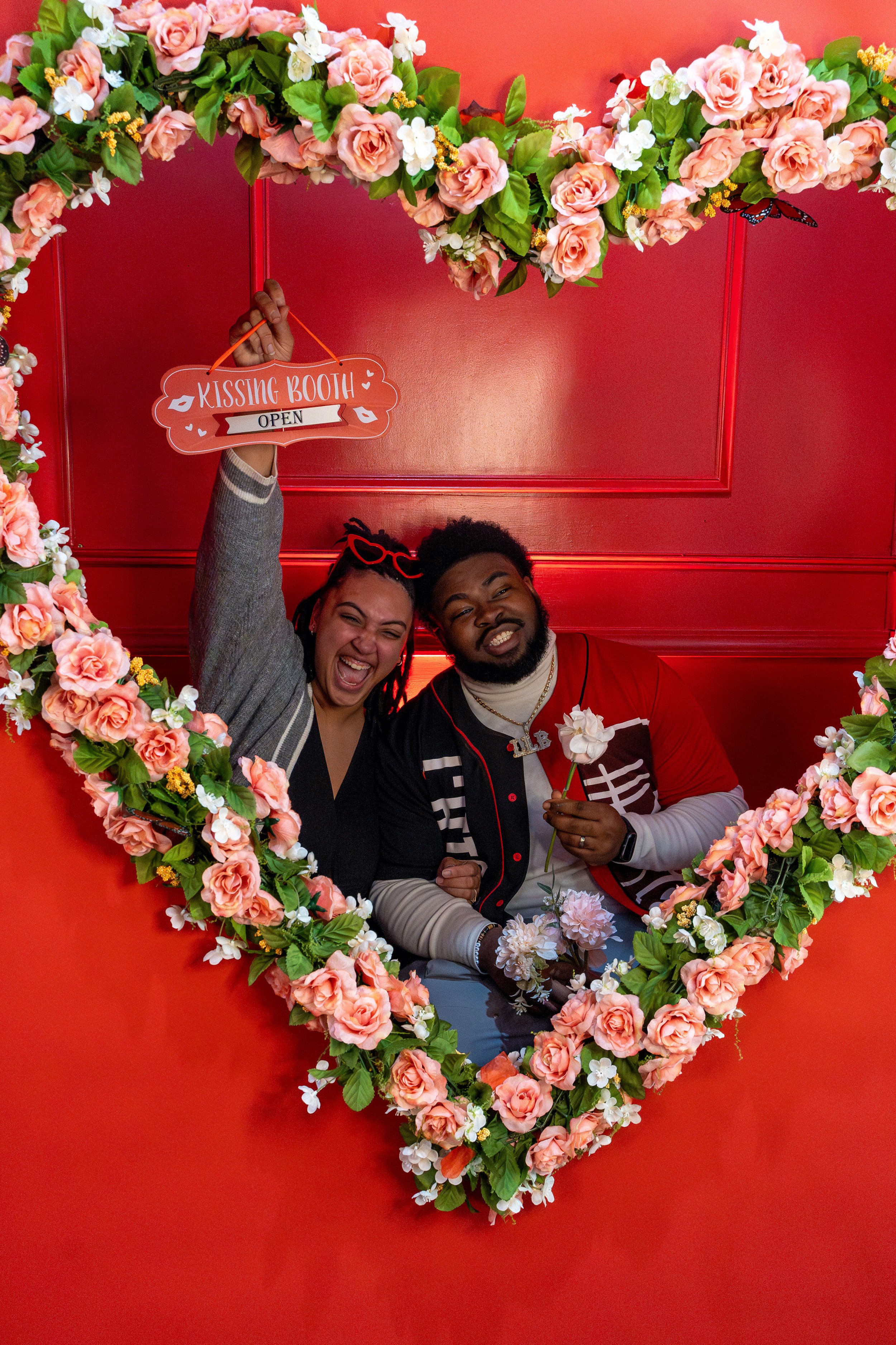 Happy couple smiling inside a heart-shaped floral frame with pink roses and white flowers, holding a sign that reads 'Kissing Booth OPEN' at a Valentine's themed event.
