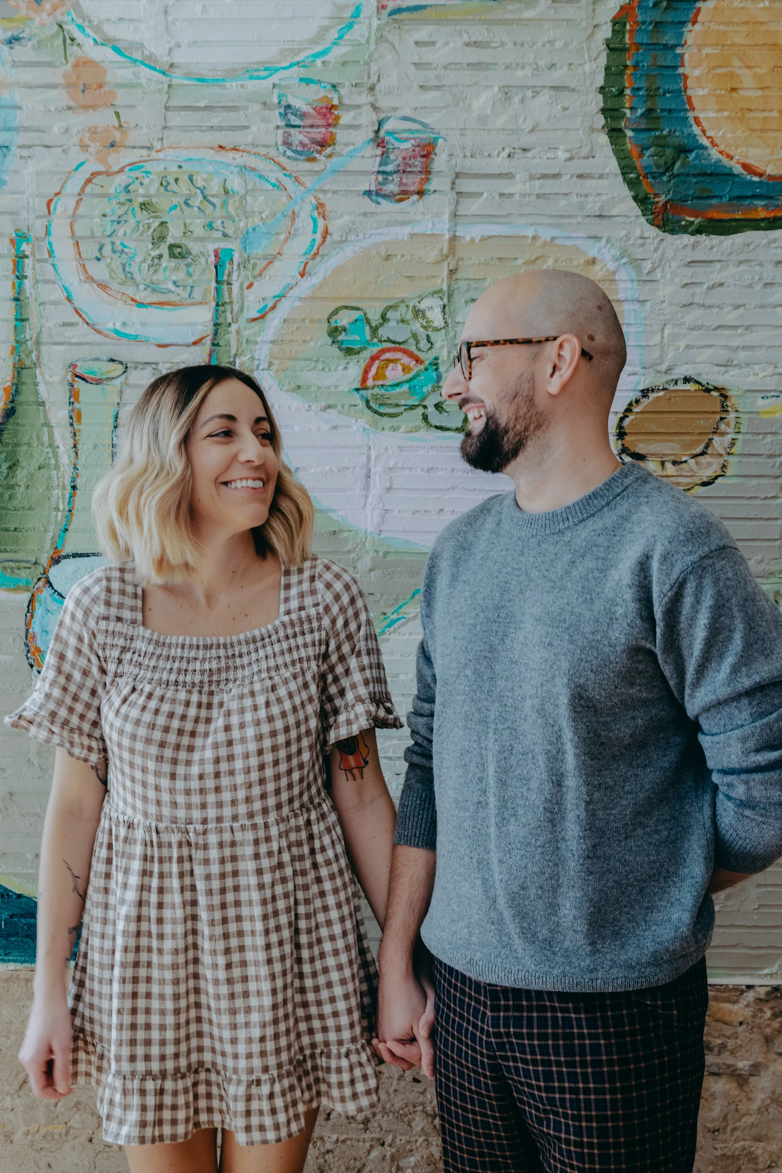A smiling couple holding hands and looking at each other in front of a colorful mural on a brick wall.