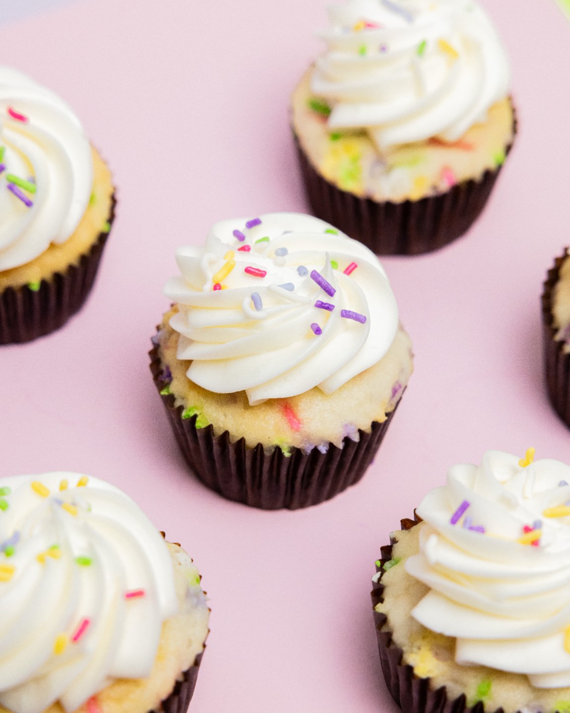 Cupcakes with white frosting and colorful sprinkles on a pink background.