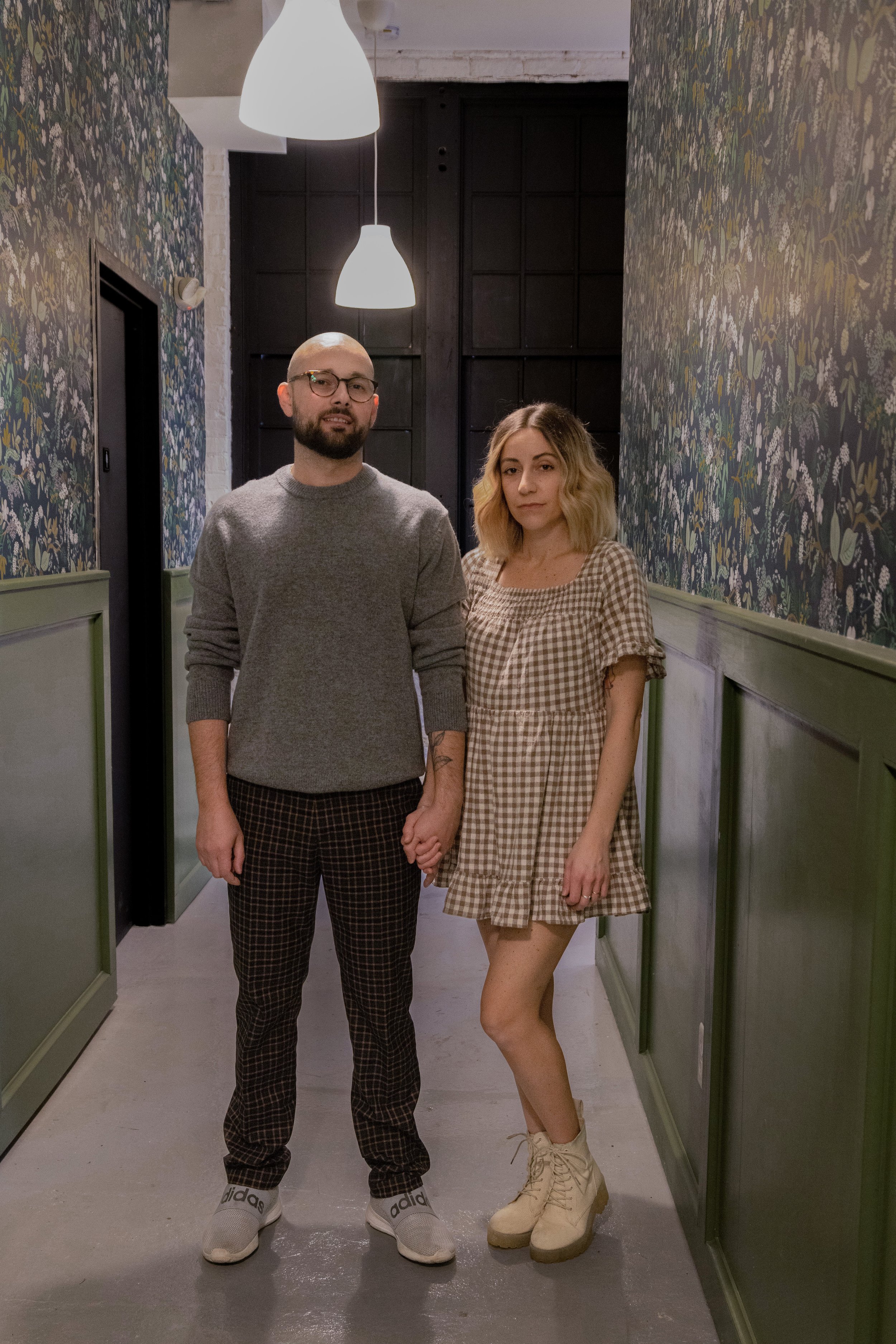 A man and woman holding hands in a hallway with floral wallpaper and green wainscoting, illuminated by hanging pendant lights.