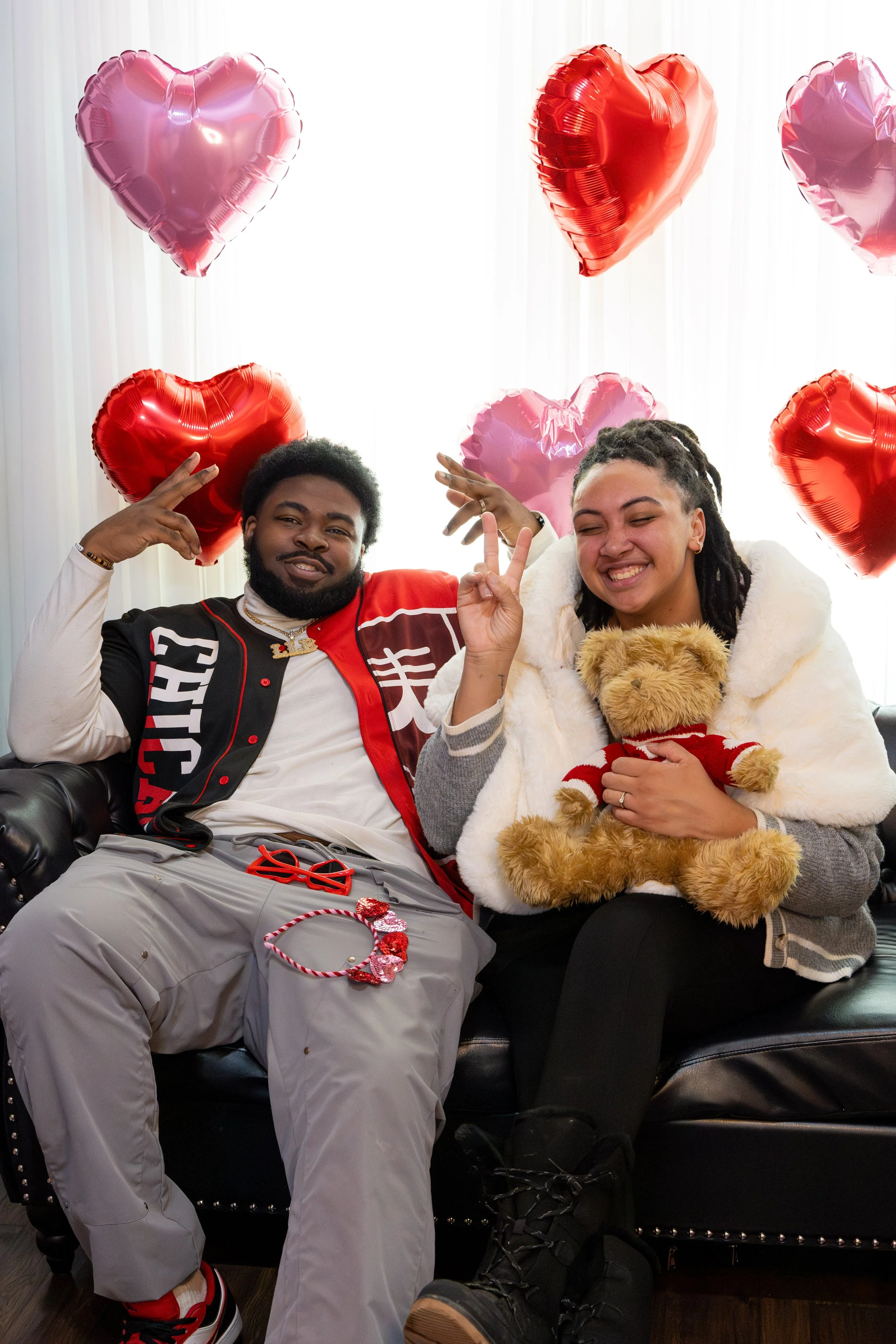 A man and a woman sitting on a black leather couch surrounded by heart-shaped balloons, with the woman holding a teddy bear.