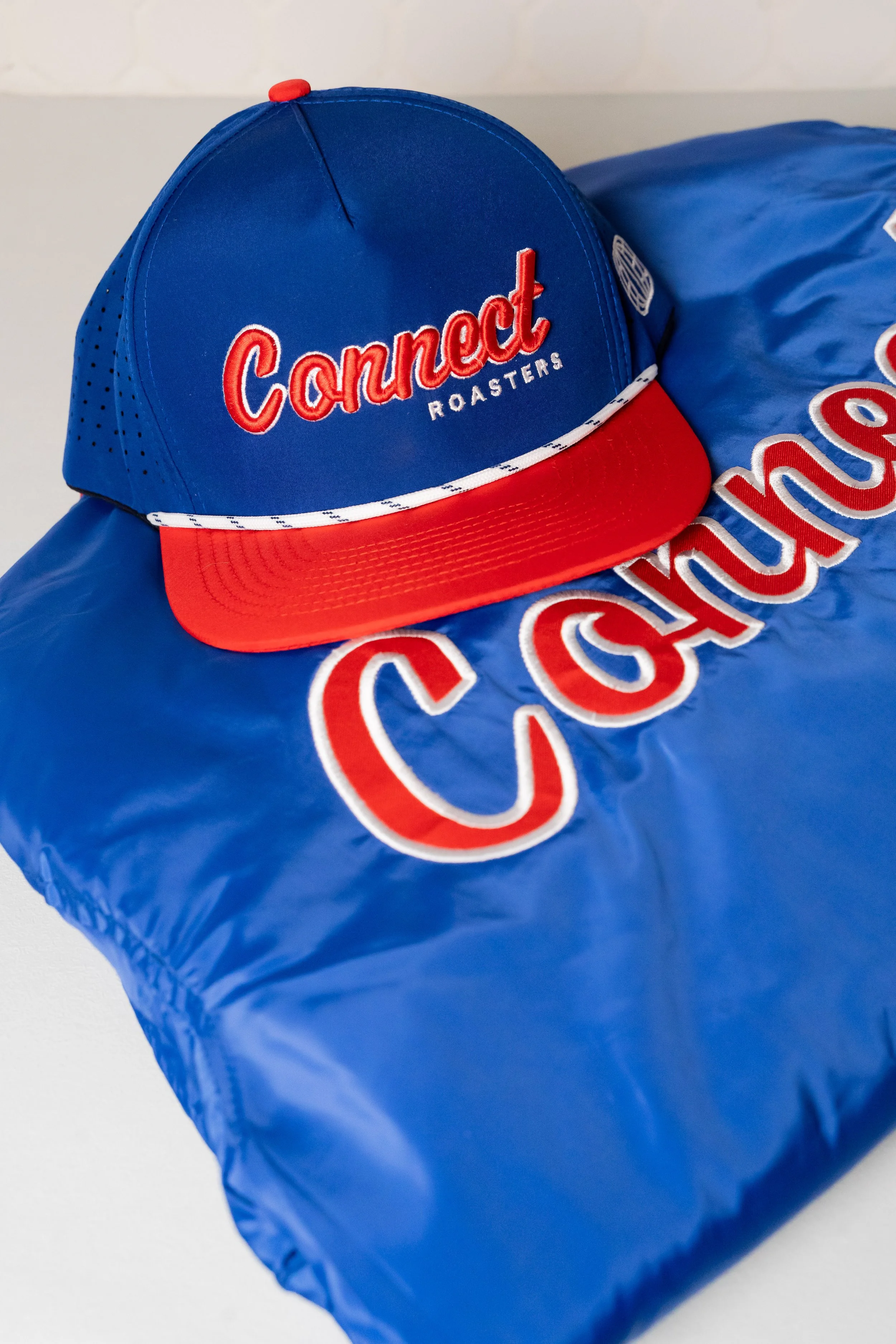 A blue baseball cap with red and white accents sitting on a blue branded bag with the words "Connect Roasters" embroidered on it.