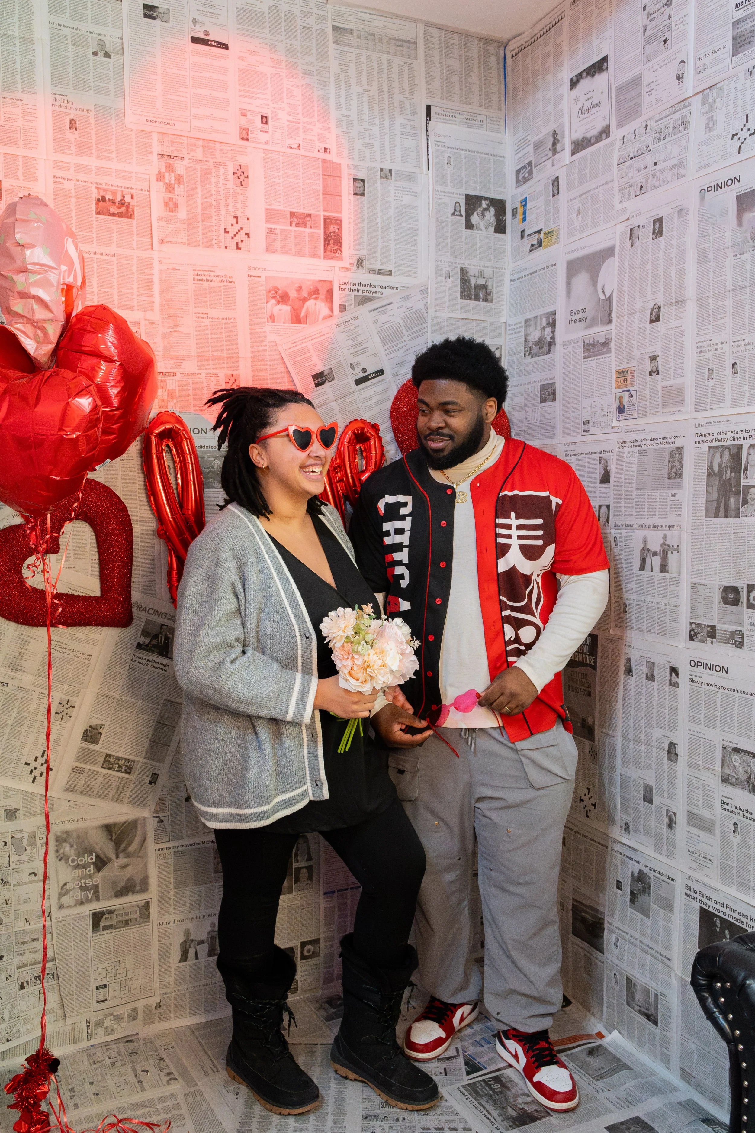 A couple at a Valentine's Day celebration, with the woman holding a bouquet of flowers and wearing heart-shaped sunglasses, and the man wearing a varsity jacket, inside a room decorated with newspapers on the walls, red balloons, and heart-shaped decorations.
