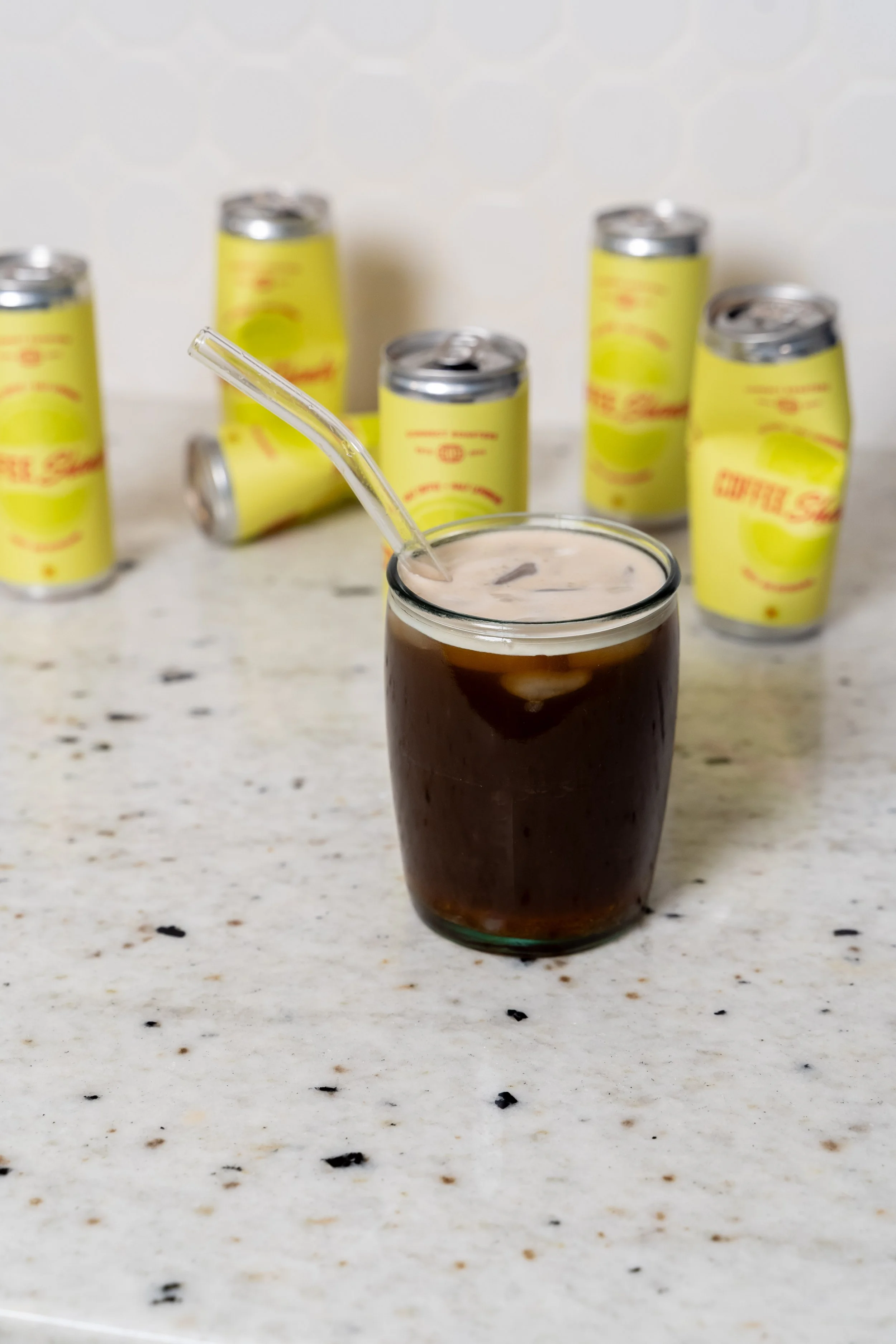 A glass of cola with ice and a straw, with multiple yellow cans in the background on a speckled white countertop.