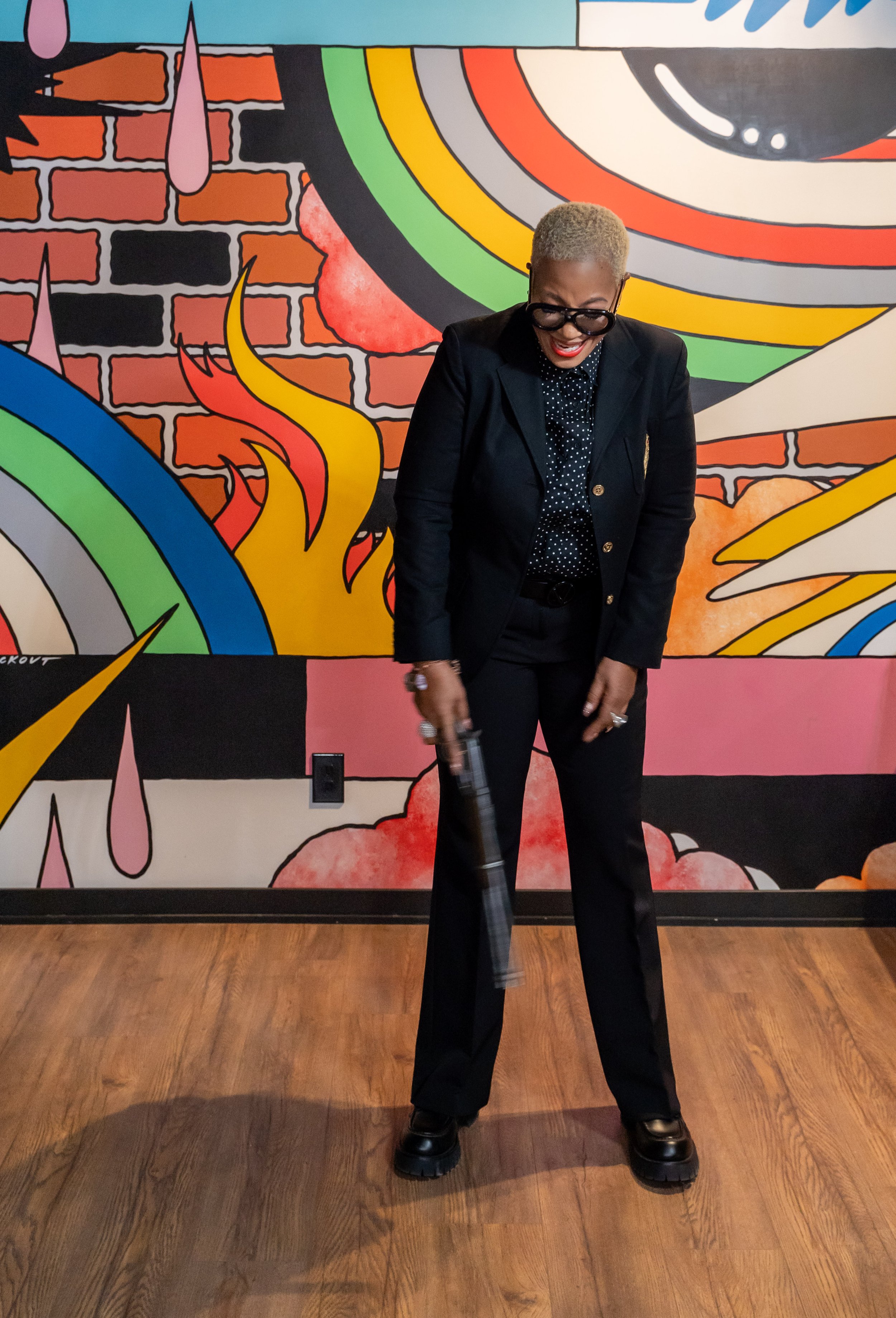 A woman with short blonde hair, glasses, and dark clothing holding a gun, standing in front of a colorful street art mural with rainbows, flames, and a brick wall.