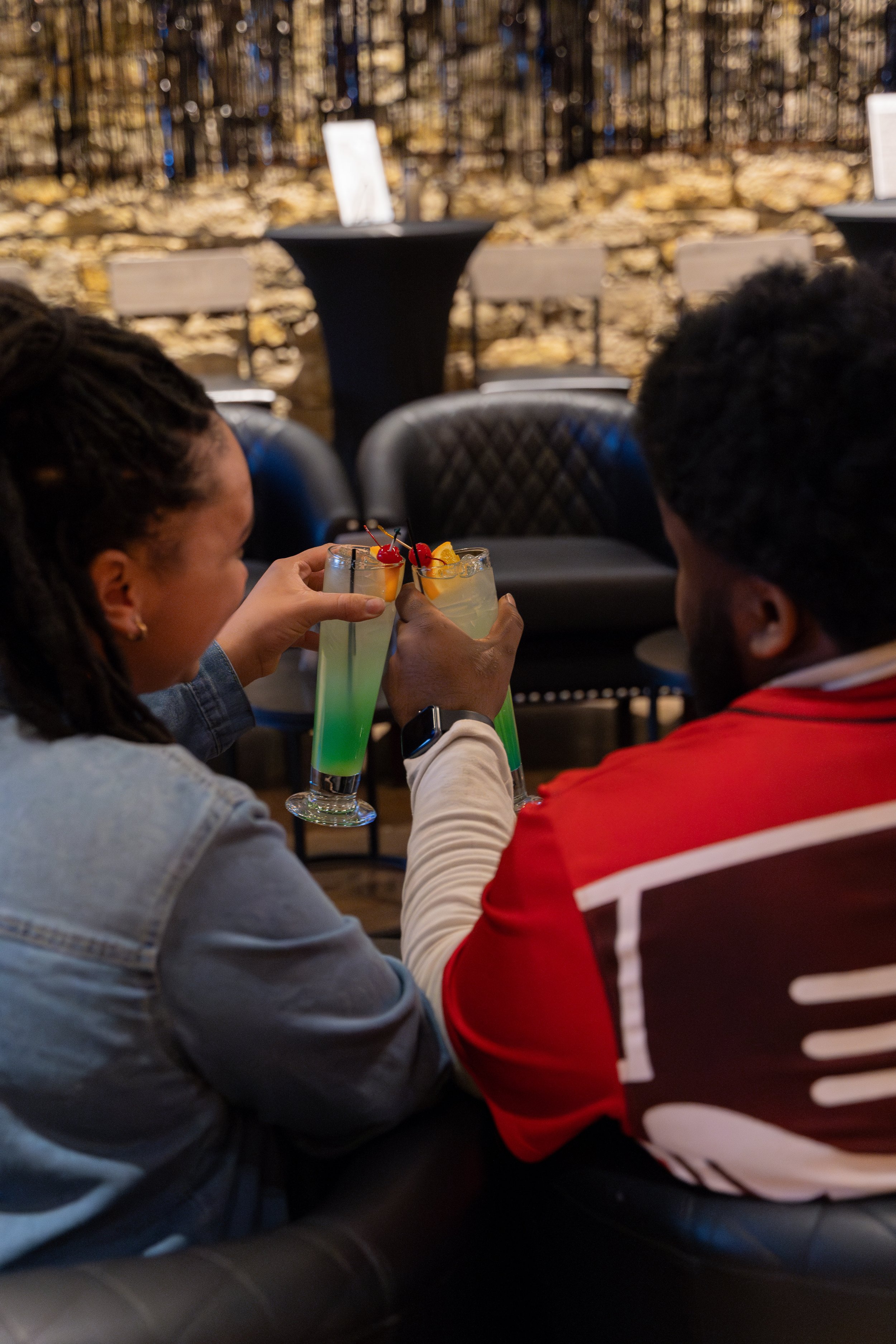 Two people clink colorful cocktails with cherries and fruit garnishes in a dimly lit lounge or bar setting.