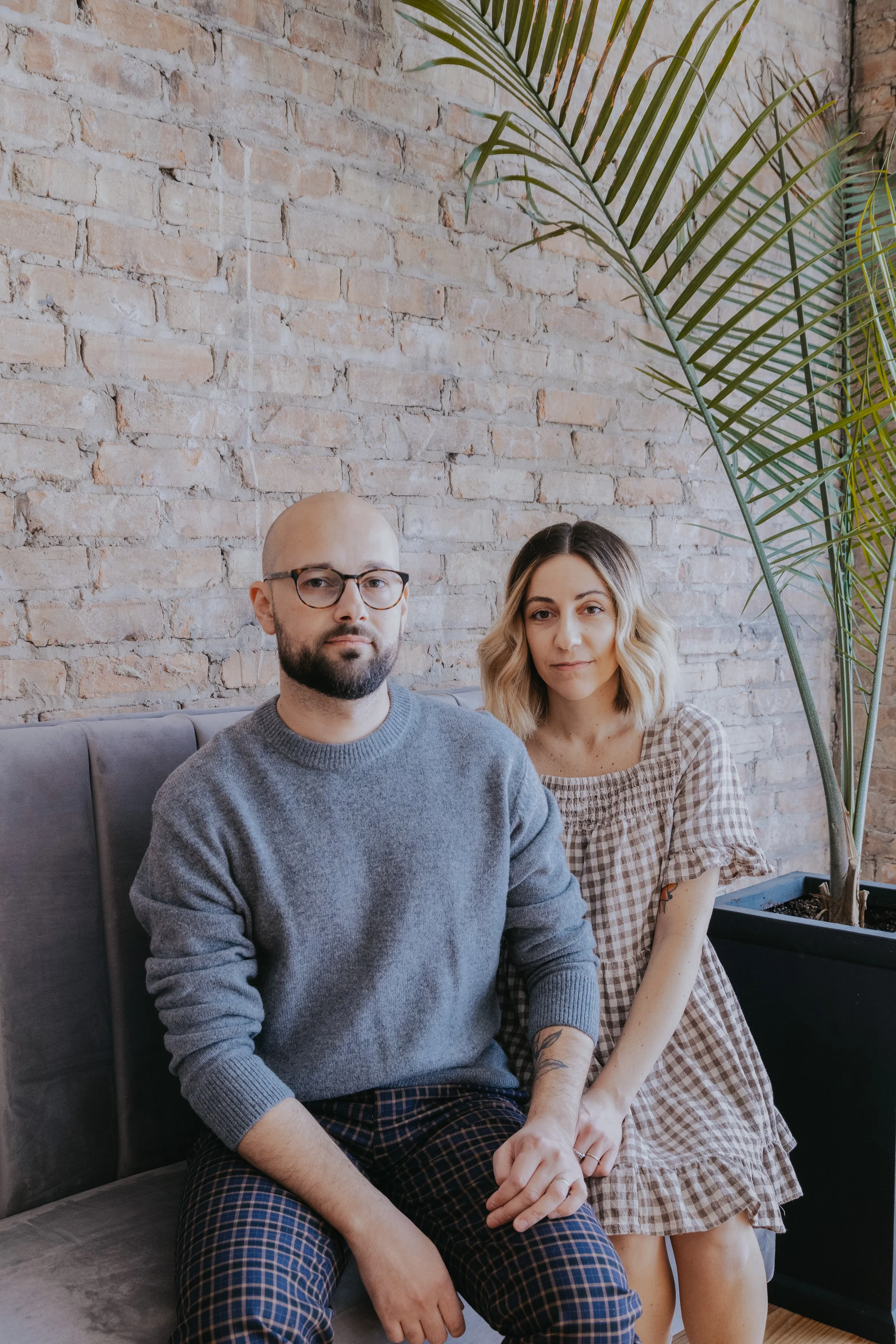 A man and woman sitting close together on a grey bench or sofa in front of a brick wall. The man has glasses, a bald head, beard, and is wearing a grey sweater and plaid pants. The woman has shoulder-length blond hair, and is wearing a beige and whit