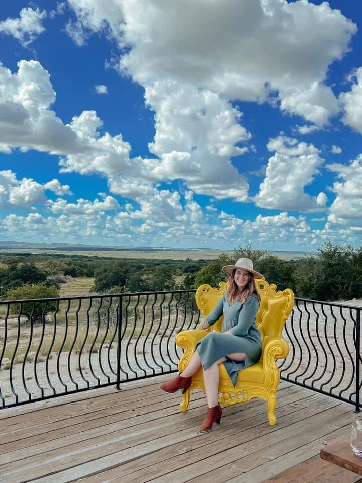 A woman sitting on a bright yellow ornate throne-like chair on a wooden deck, wearing a blue dress, a wide-brimmed hat, and brown ankle boots, with a landscape of green bushes and trees and a blue sky with puffy white clouds in the background.