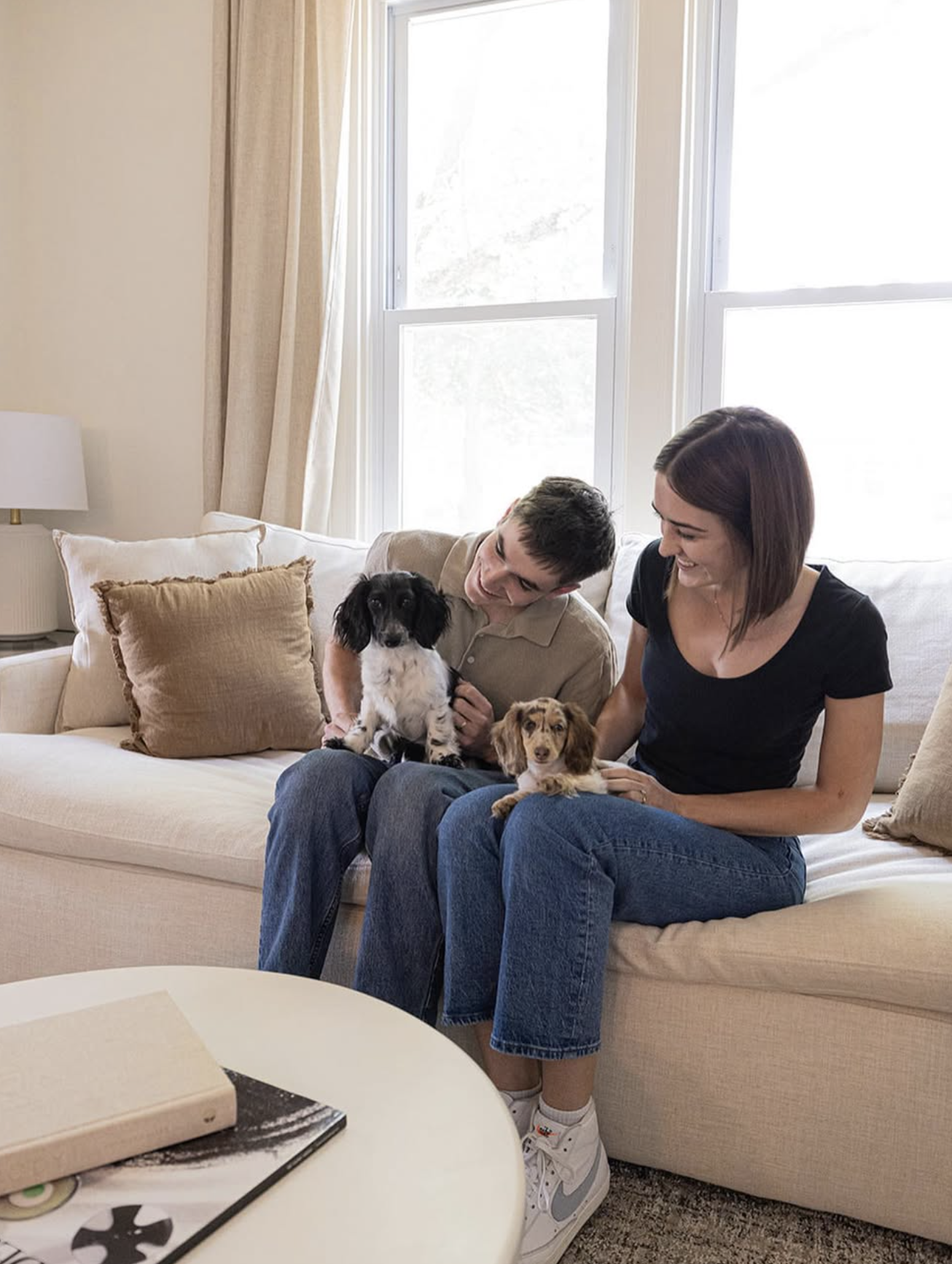 A happy couple sitting on a beige couch with two dogs, smiling and interacting in a bright living room with large windows and beige curtains.