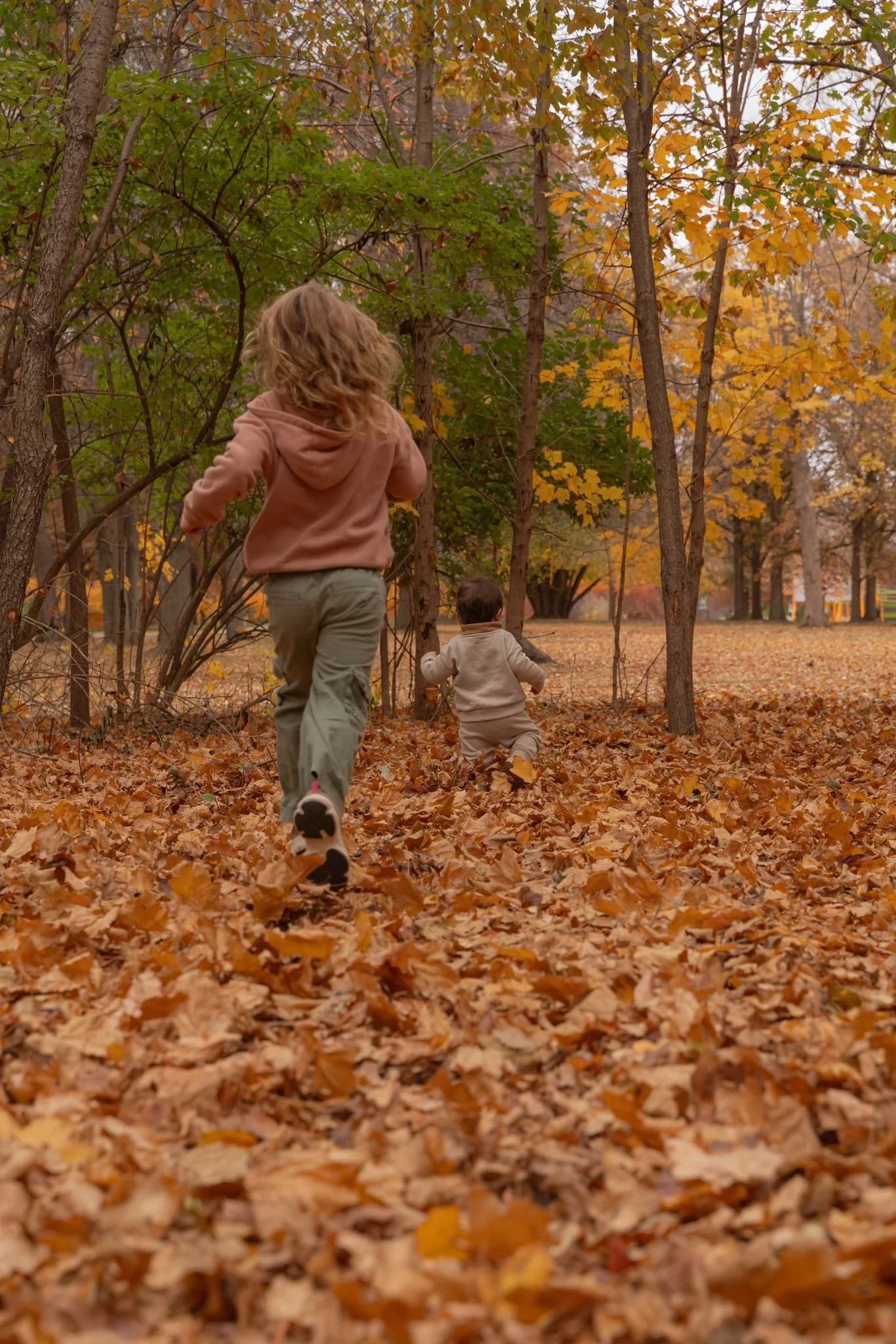 A girl and a toddler running through an autumn forest covered with fallen orange and brown leaves, surrounded by trees with yellow and orange foliage.