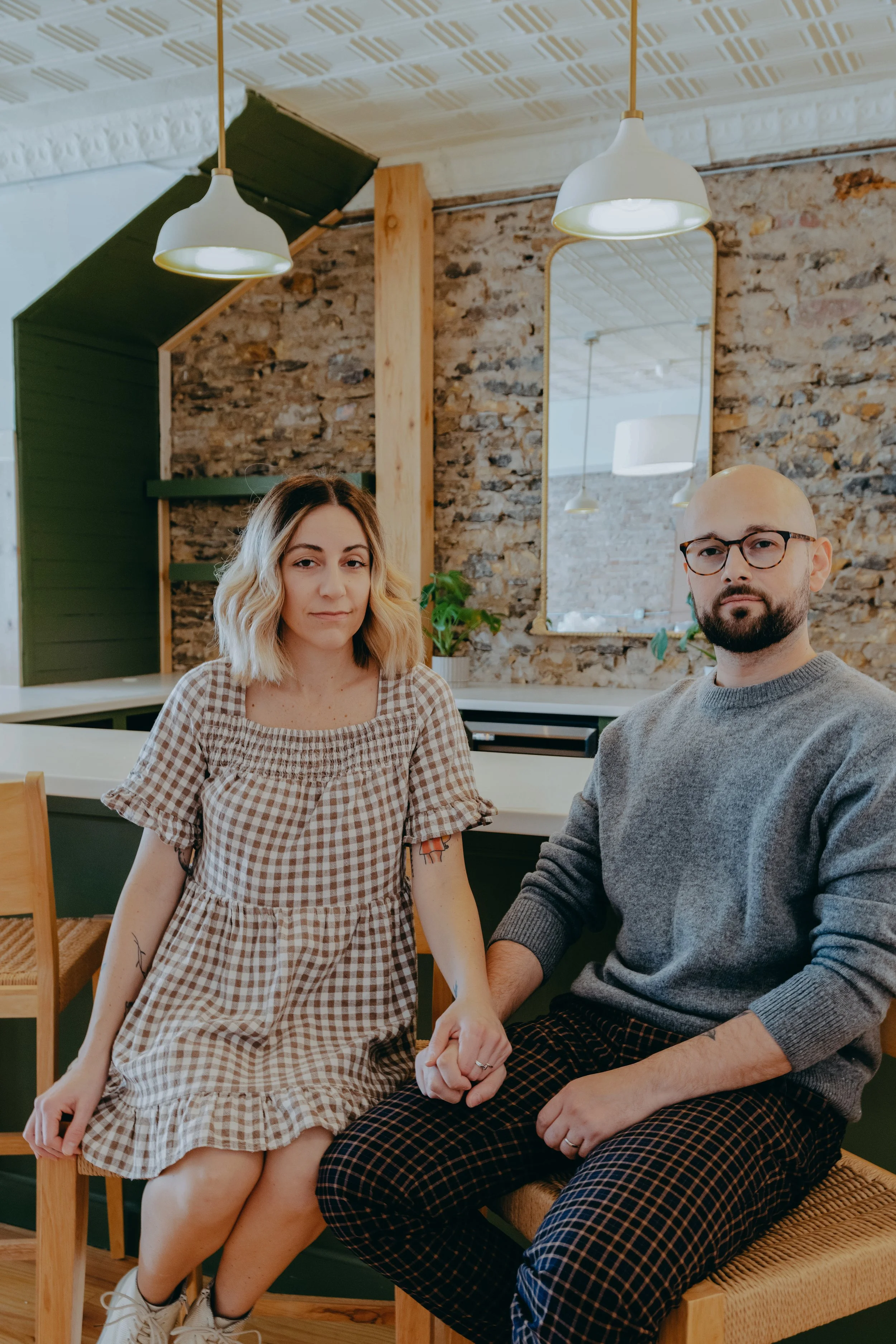 A man and a woman holding hands sit in a cozy room with a brick wall and a mirror behind them. The woman is wearing a gingham dress, and the man is wearing a gray sweater and plaid pants. Soft lighting comes from hanging ceiling lamps.