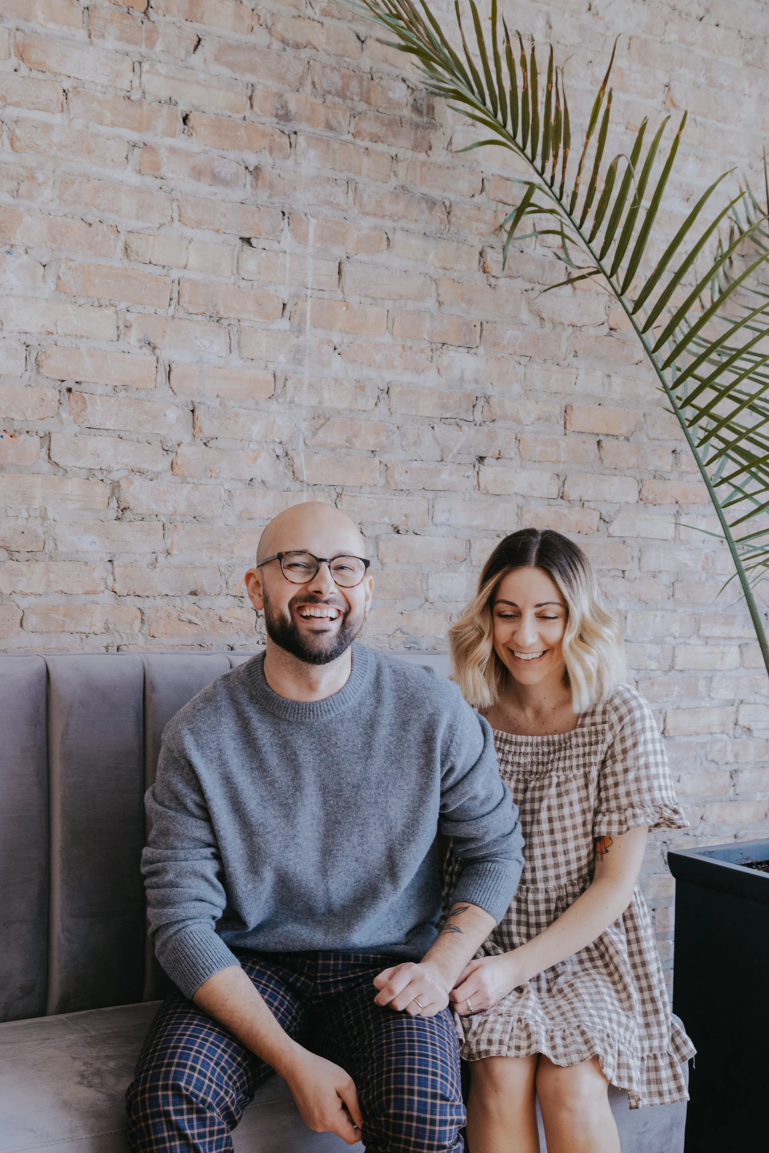 A man with glasses and a woman with blonde hair sitting together on a gray sofa, smiling and laughing. They are in a room with an exposed brick wall and a large green plant.