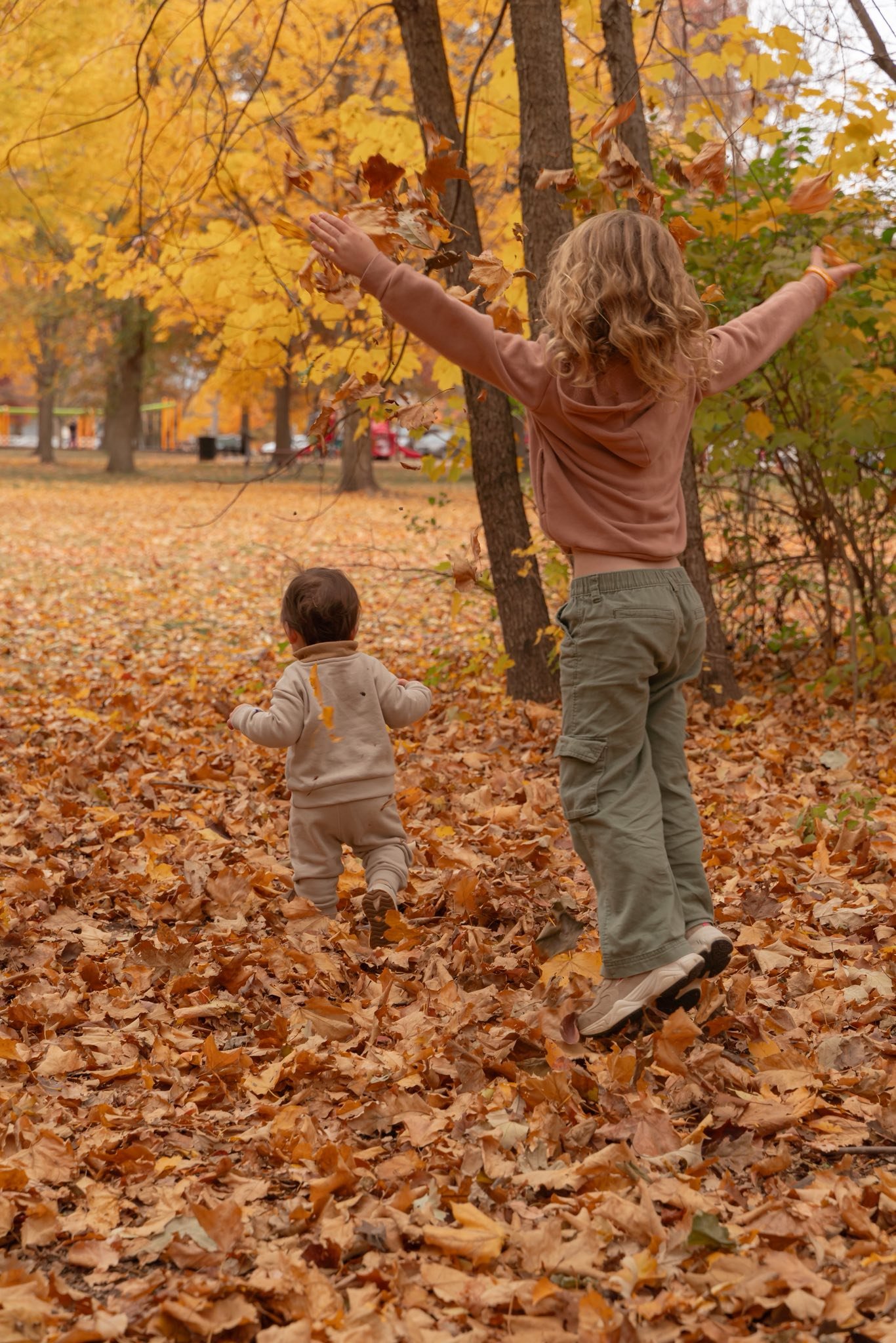 A woman and a small child playing in a park filled with fallen autumn leaves. The woman is tossing leaves into the air, while the child runs through the leaves, surrounded by trees with yellow and orange foliage.
