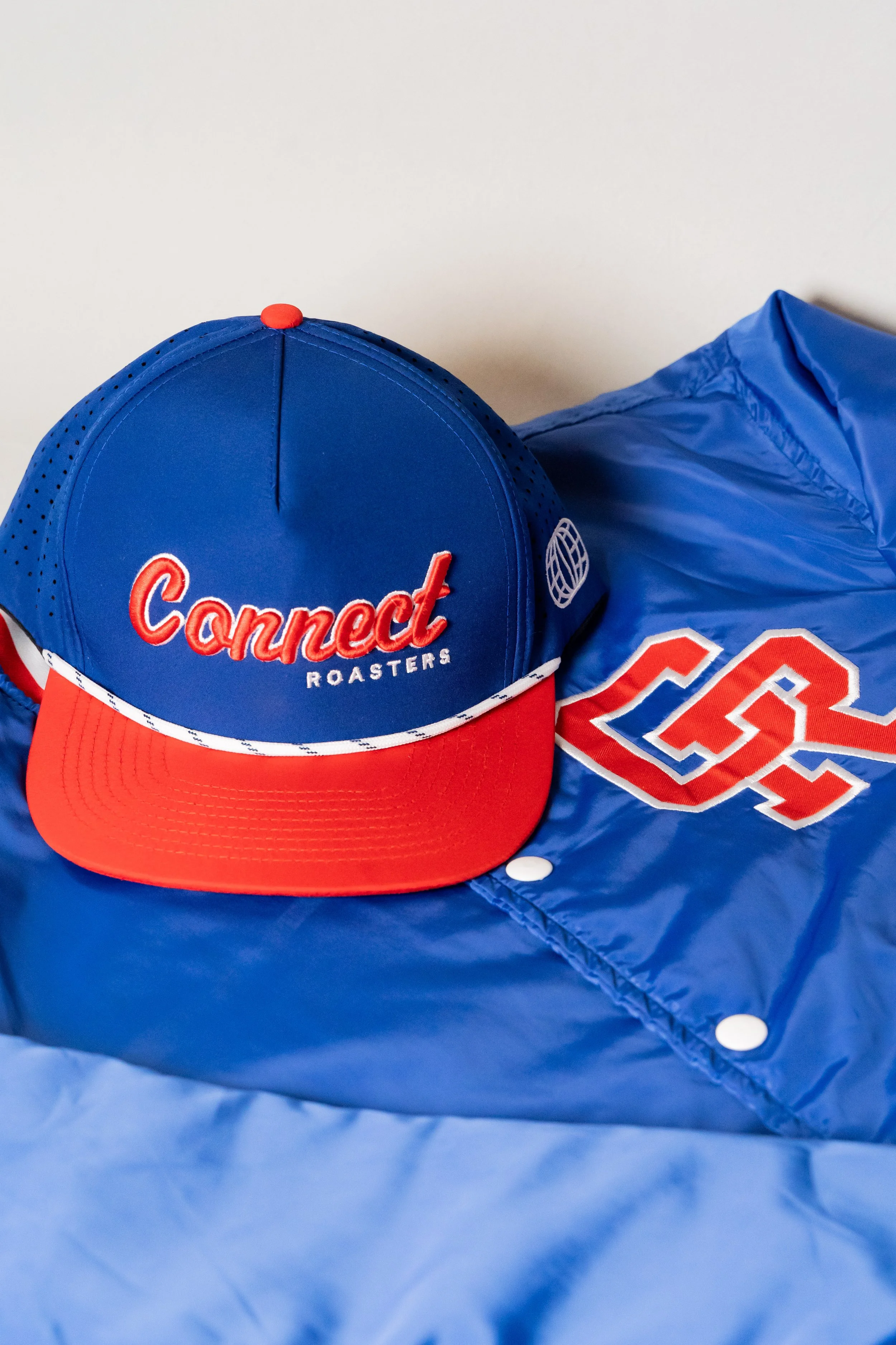 A blue and red baseball cap with 'Connect Roasters' embroidered on the front, placed on a blue jacket with a white and red logo.