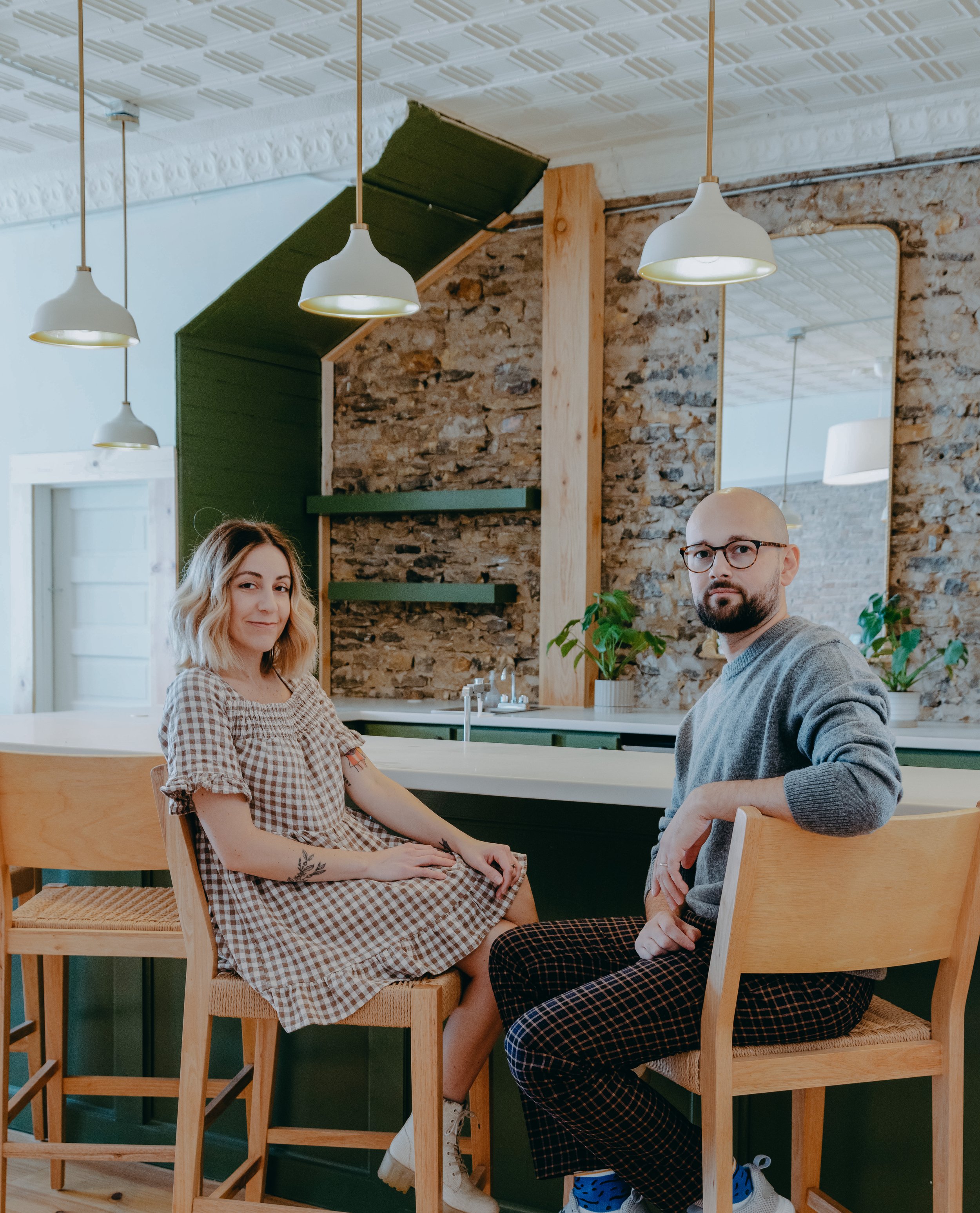 A woman and a man sitting at a kitchen island in a modern kitchen with exposed brick wall, green accents, and pendant lighting.