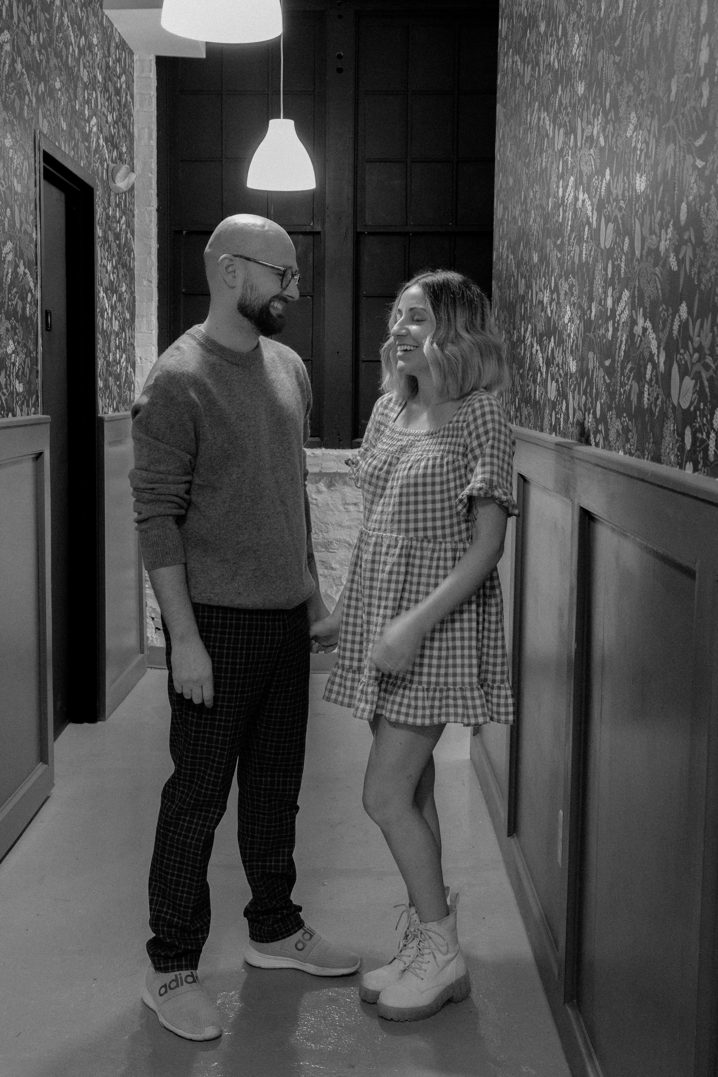 A man and woman smiling and holding hands in a cozy indoor setting with floral wallpaper and warm lighting.