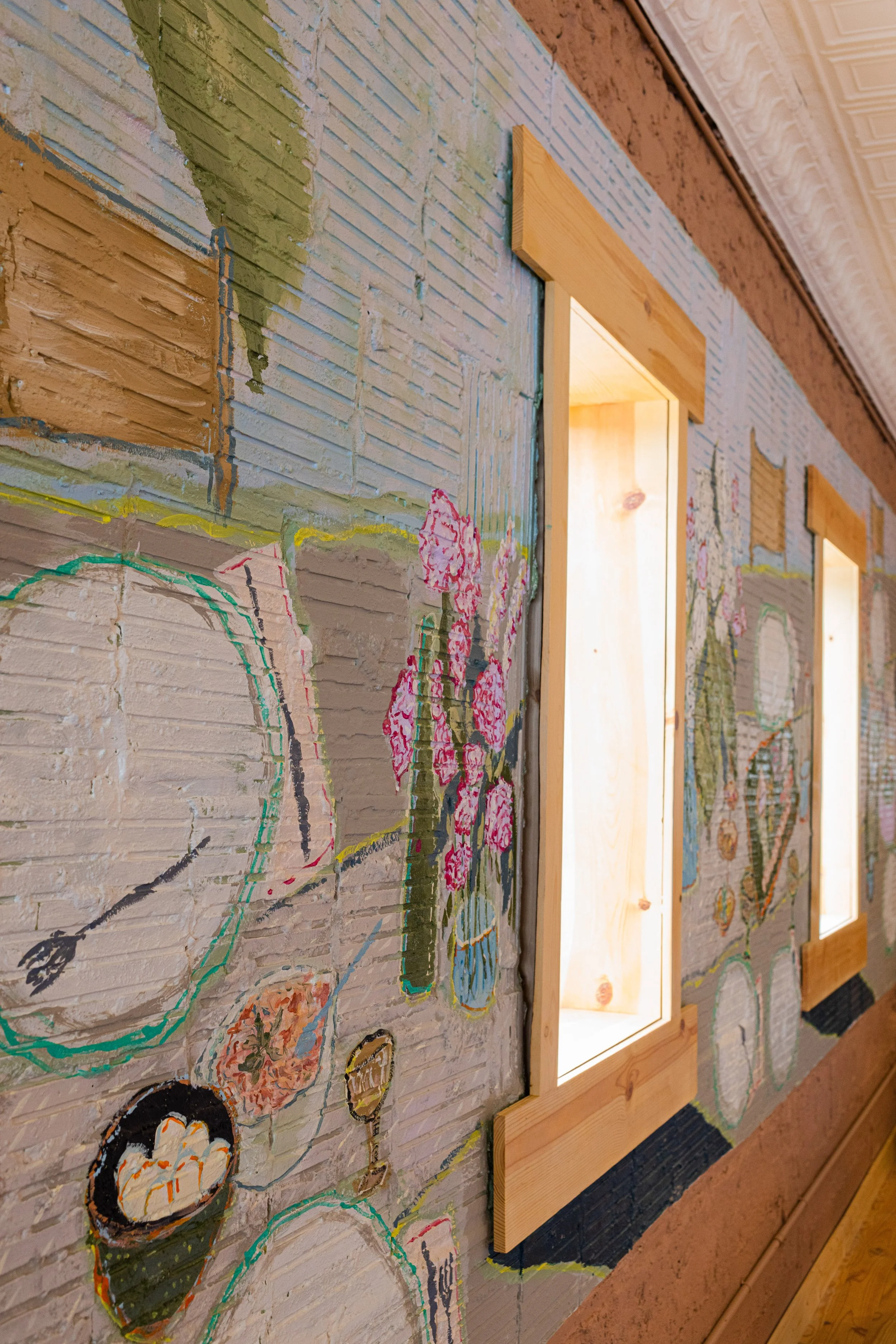 Colorful mural of a still life scene with a bowl of rice, glass of wine, and pink flowers painted on a brick wall, with wooden window frames and illuminated interior lighting.
