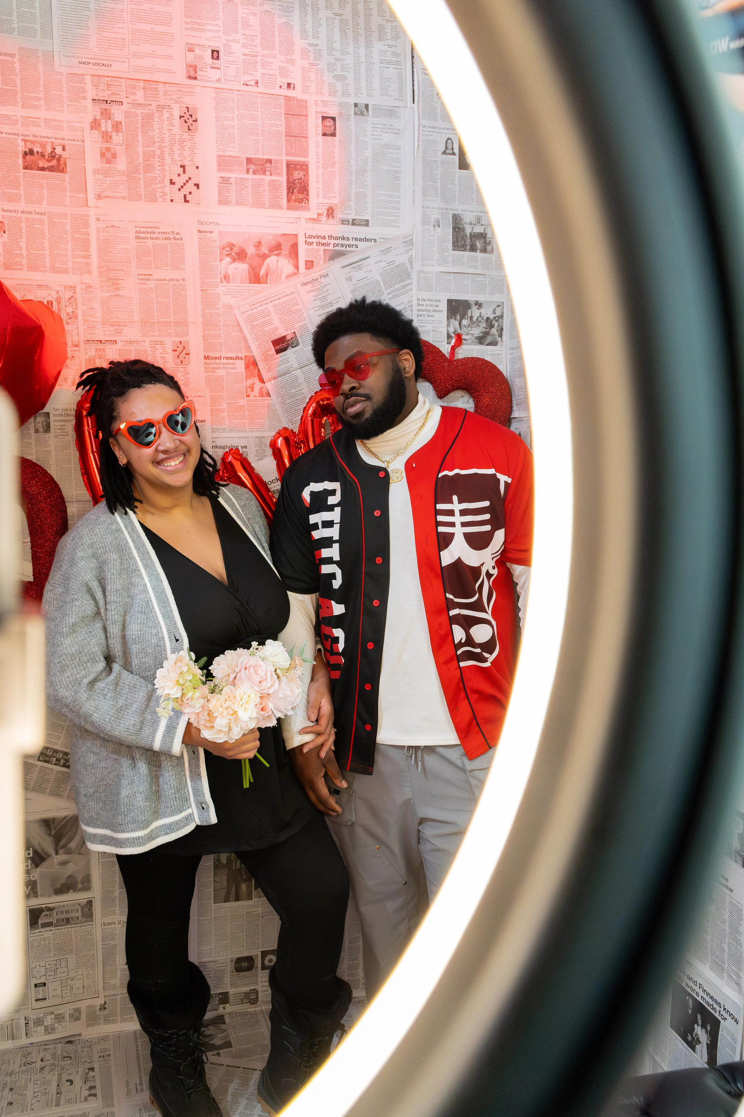 A couple posing in a photo booth, with a woman holding a bouquet of flowers and a man standing beside her, both wearing colorful, trendy outfits and accessories, with a decorated backdrop of newspapers and Valentine's Day-themed decorations.