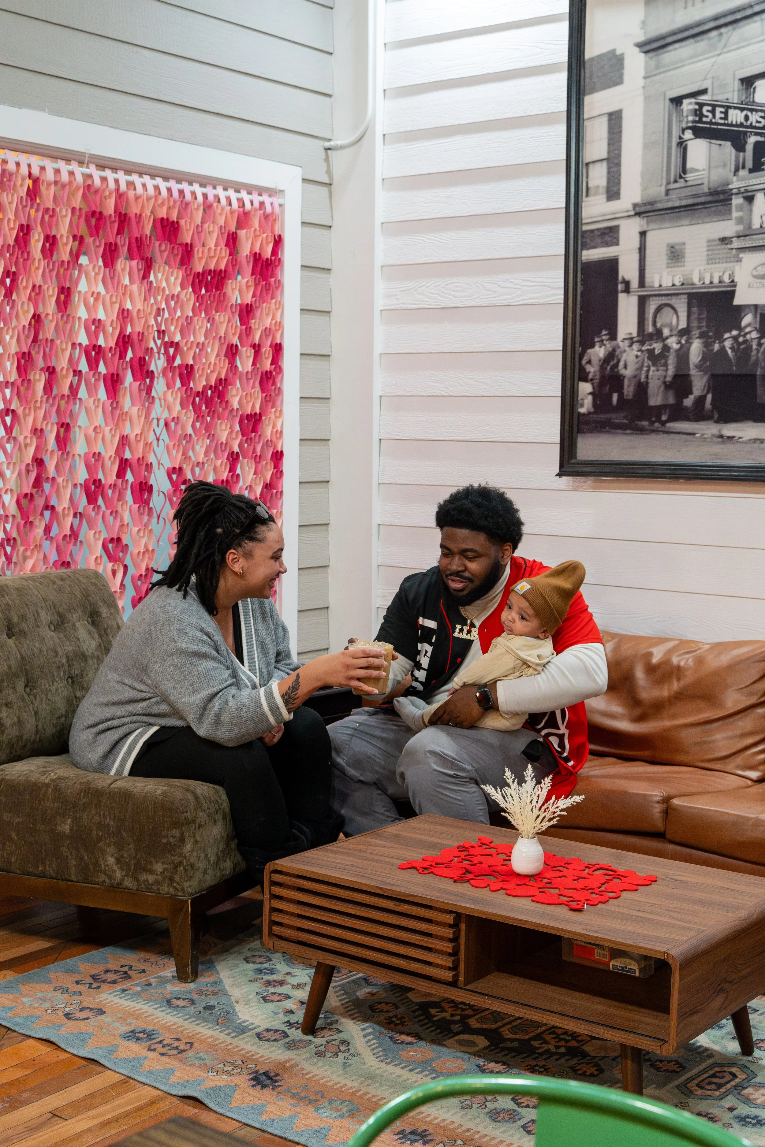 A family of three sitting on a couch in a cozy living room, with the woman smiling and holding a drink, the man holding a baby and looking at her, and the baby wearing a warm hat.