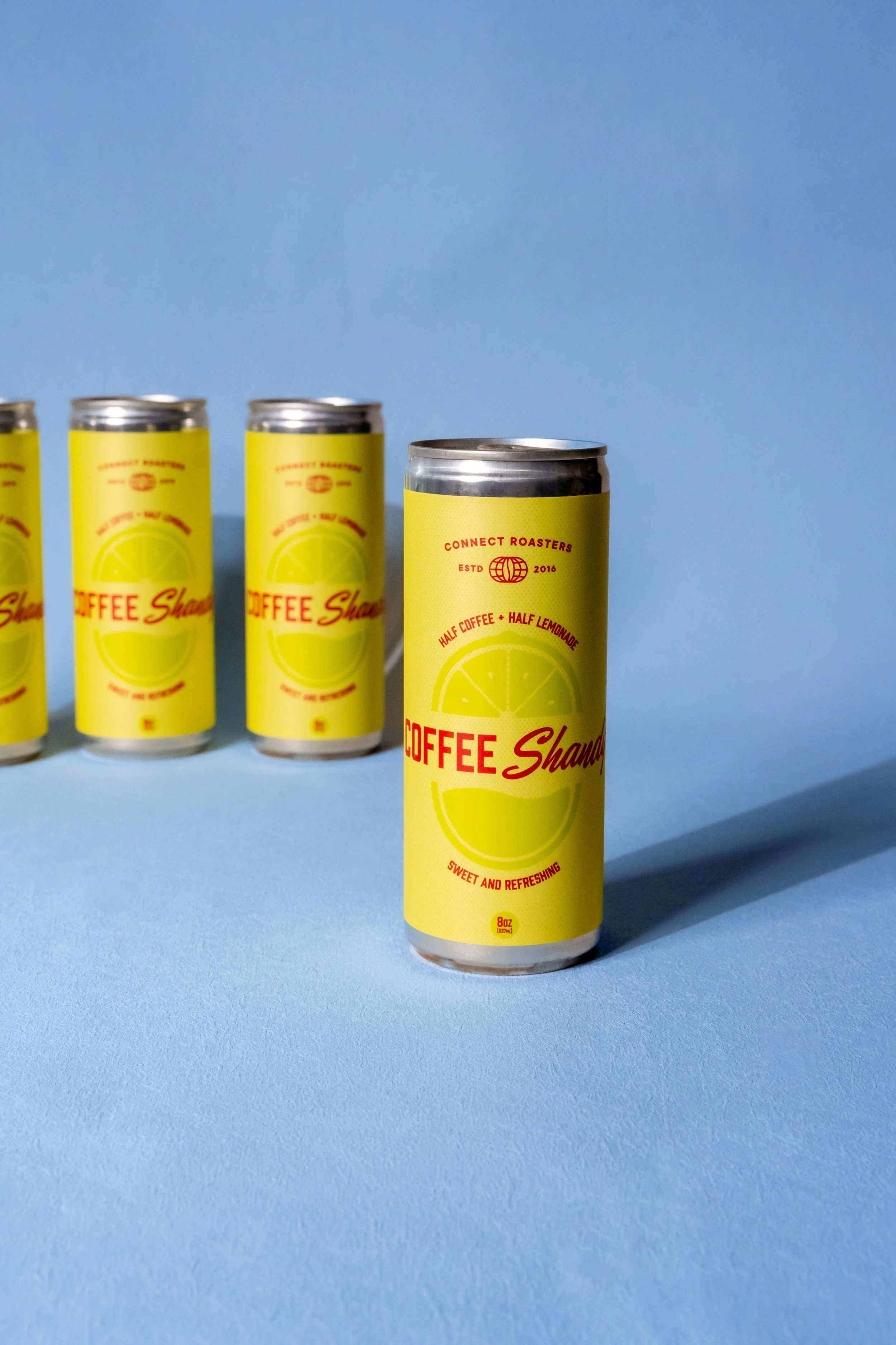 Four yellow cans of coffee lemonade beverage with red text, standing on a light blue surface against a darker blue background, with one can in the foreground and the others slightly blurred in the background.