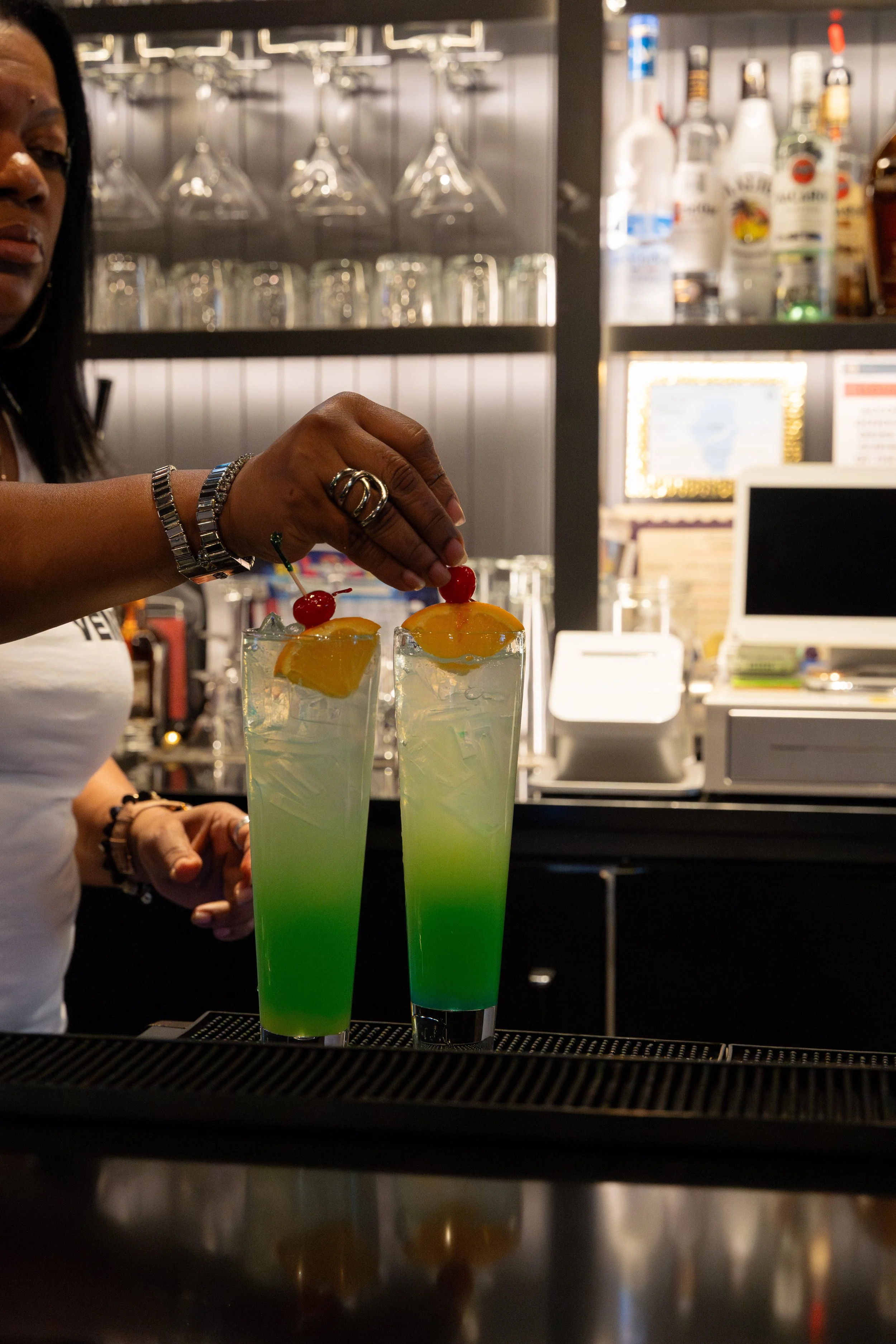 A bartender garnishing two tall, green and white layered cocktails with cherries and orange slices at a bar.