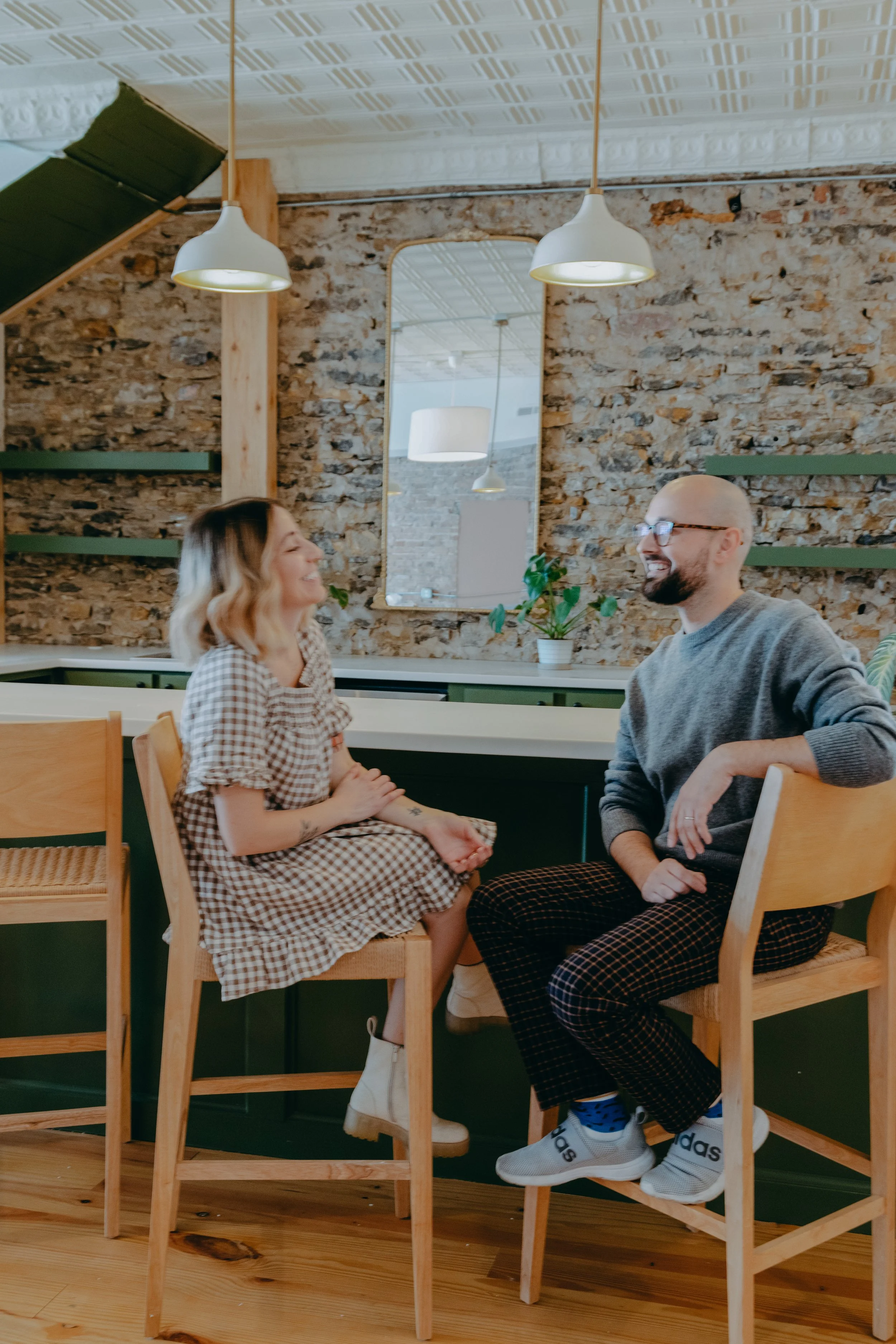 A woman and a man sitting on high chairs at a kitchen counter, smiling and engaging in conversation in a cozy, modern kitchen with a brick wall, hanging pendant lights, and a large mirror.