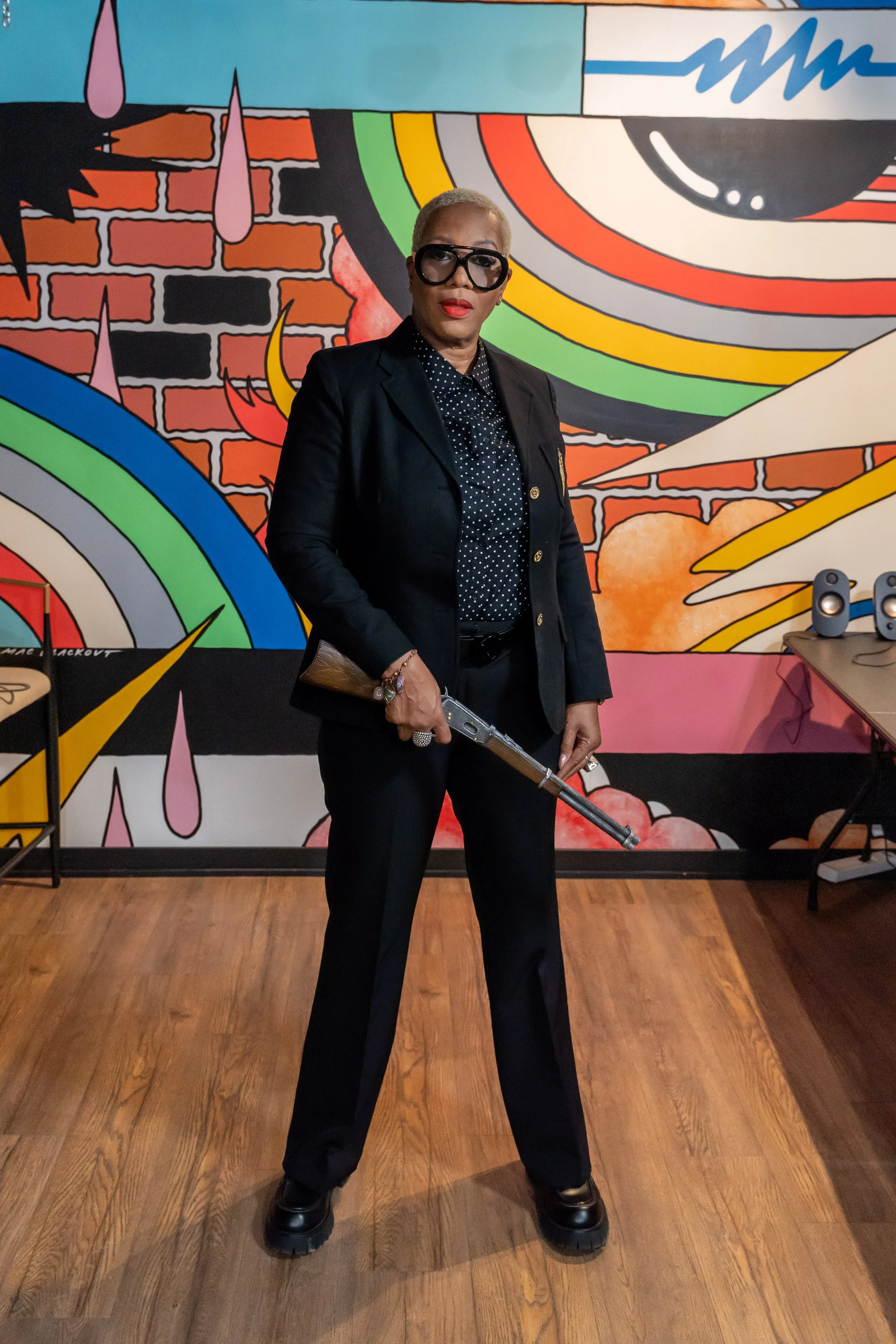 A woman dressed in a black suit and glasses standing with a rifle in front of a colorful graffiti mural featuring rainbows, clouds, and abstract designs.