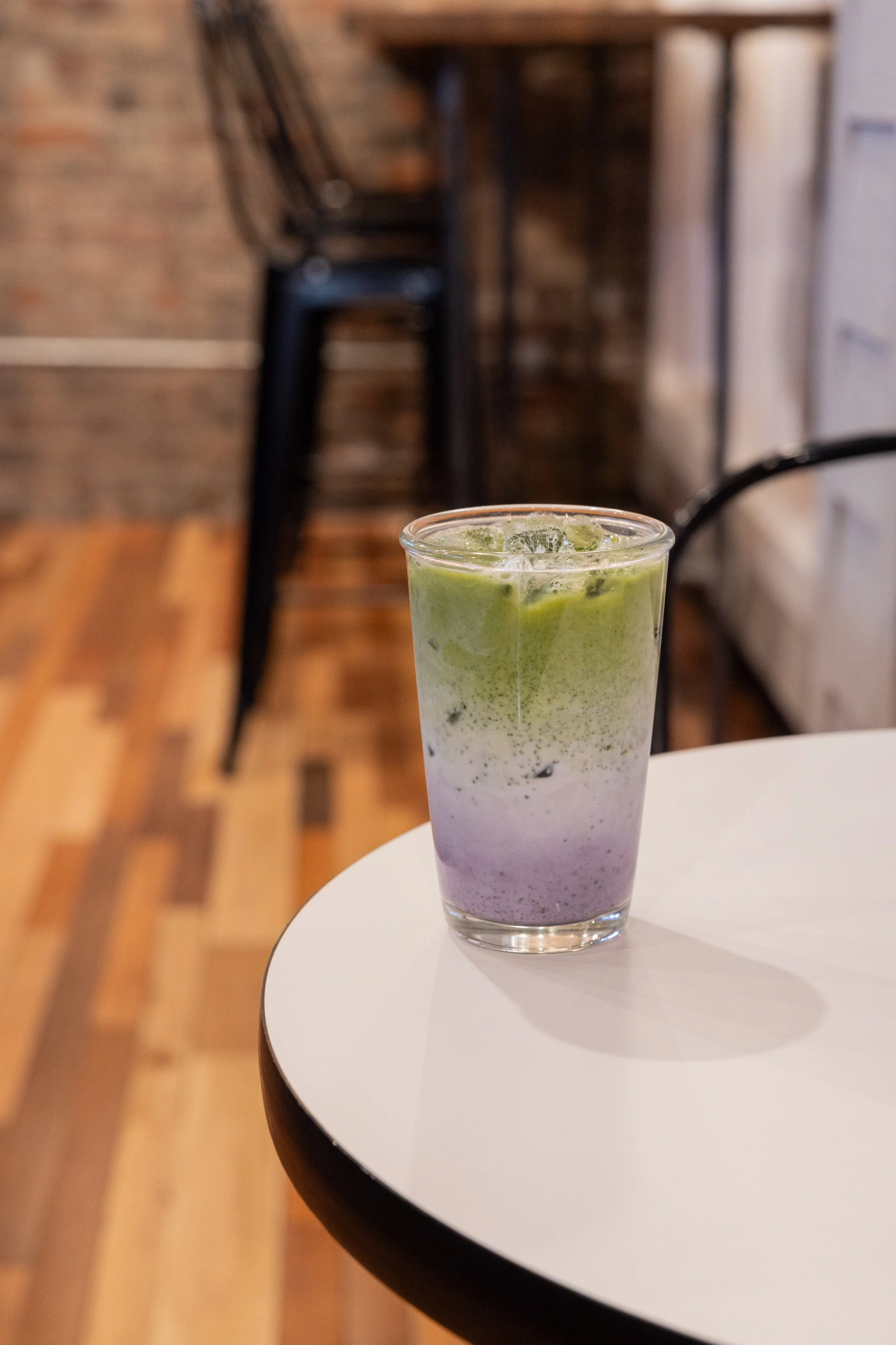 A multi-layered iced matcha latte with a purple and green gradient, served in a clear glass on a white table, in a cafe setting.