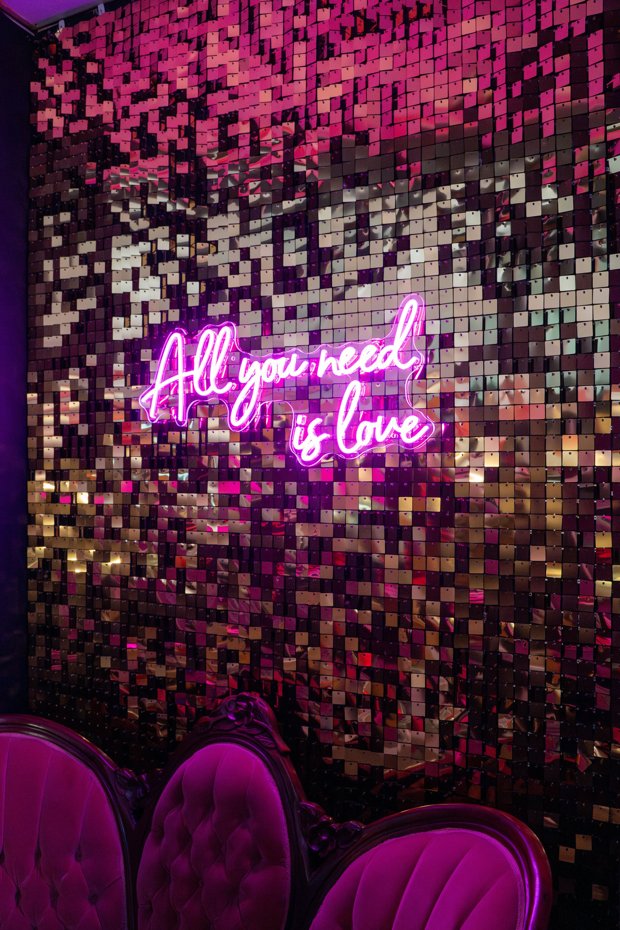Neon pink sign that says 'All you need is love' hanging on a reflective mosaic wall, with purple velvet chairs in the foreground.