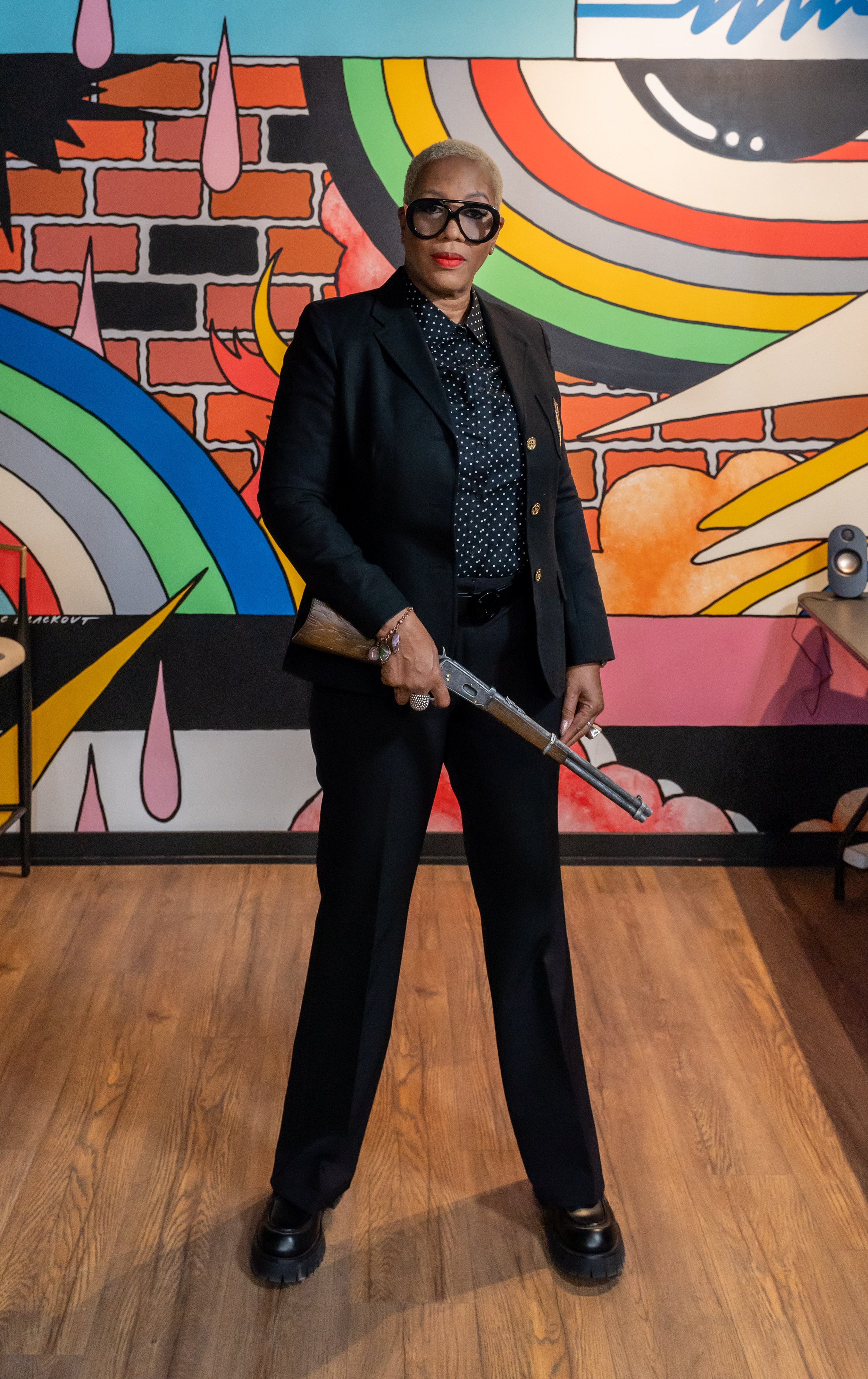 Woman with short blonde hair, wearing large black glasses, dressed in a dark suit and polka dot shirt, holding a rifle, standing in front of a colorful graffiti mural with rainbows and clouds.