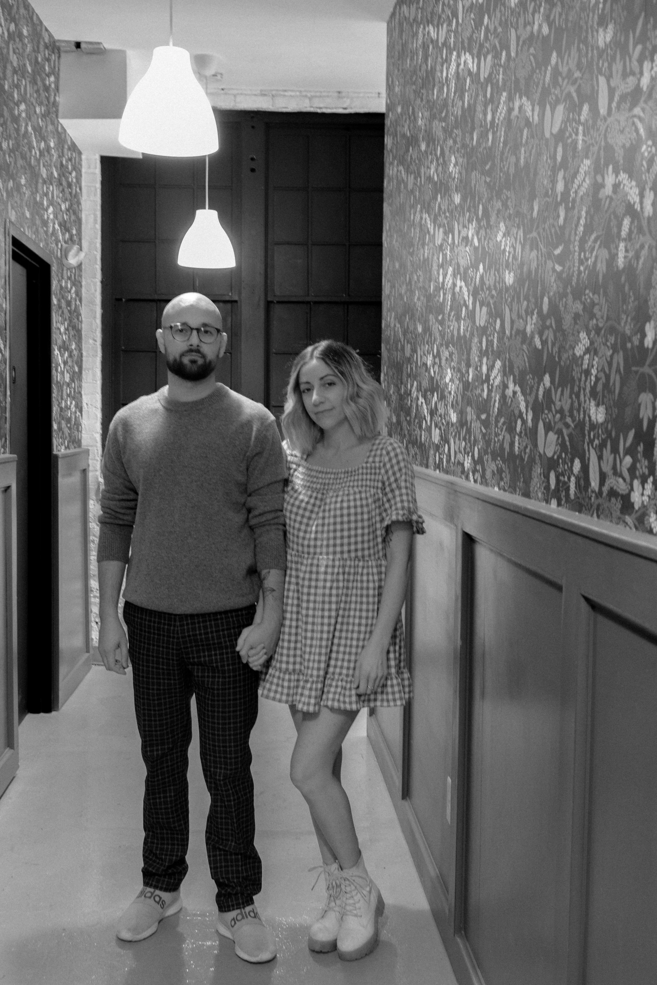 A man and woman holding hands in a hallway, both wearing glasses. The man wears a sweater and checkered pants, the woman wears a checkered dress and boots. The hallway has paneling and floral wallpaper, with pendant lights hanging from the ceiling.