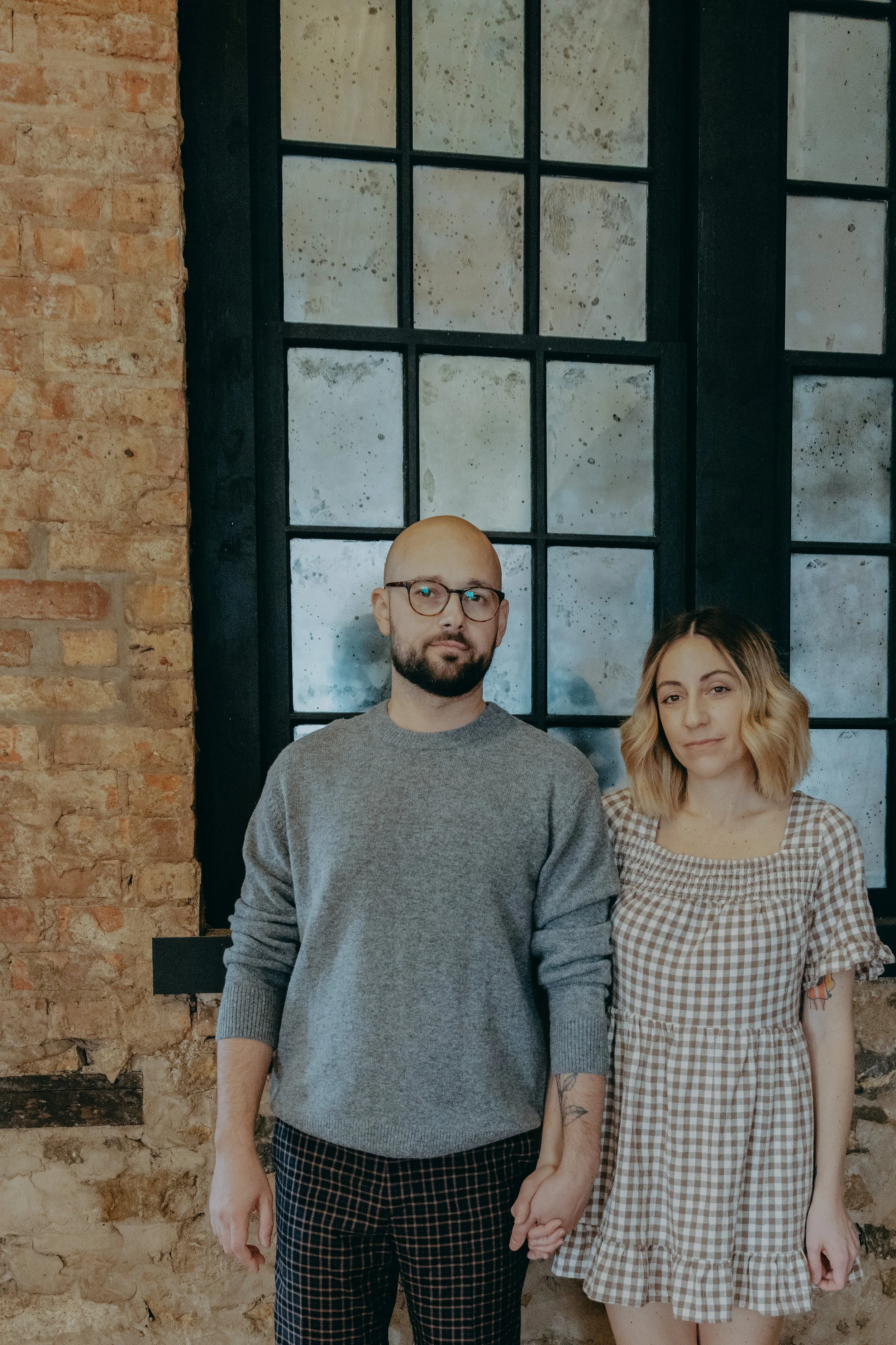 A man and woman holding hands indoors in front of a large industrial-style window, with exposed brick wall on the left side.